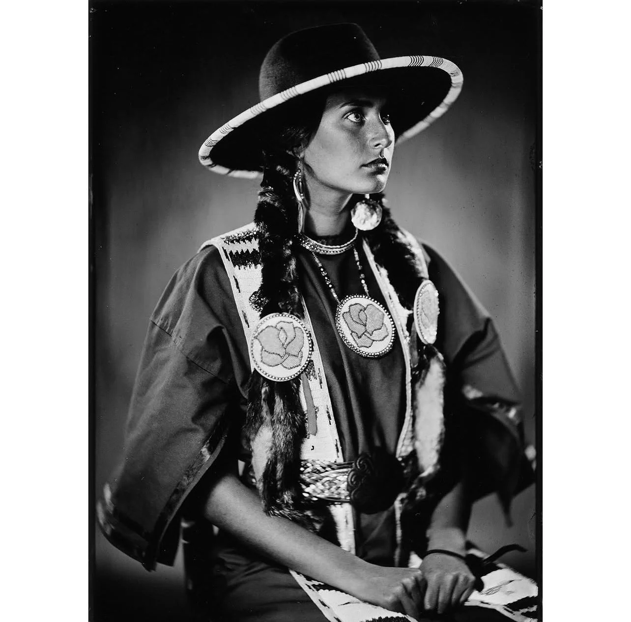 A black-and-white photo of a Native American woman dressed in traditional attire, wearing a large hat with a wide brim, decorated necklaces, earrings, and a patterned shawl with medallions, sitting with her hands resting on her lap.
