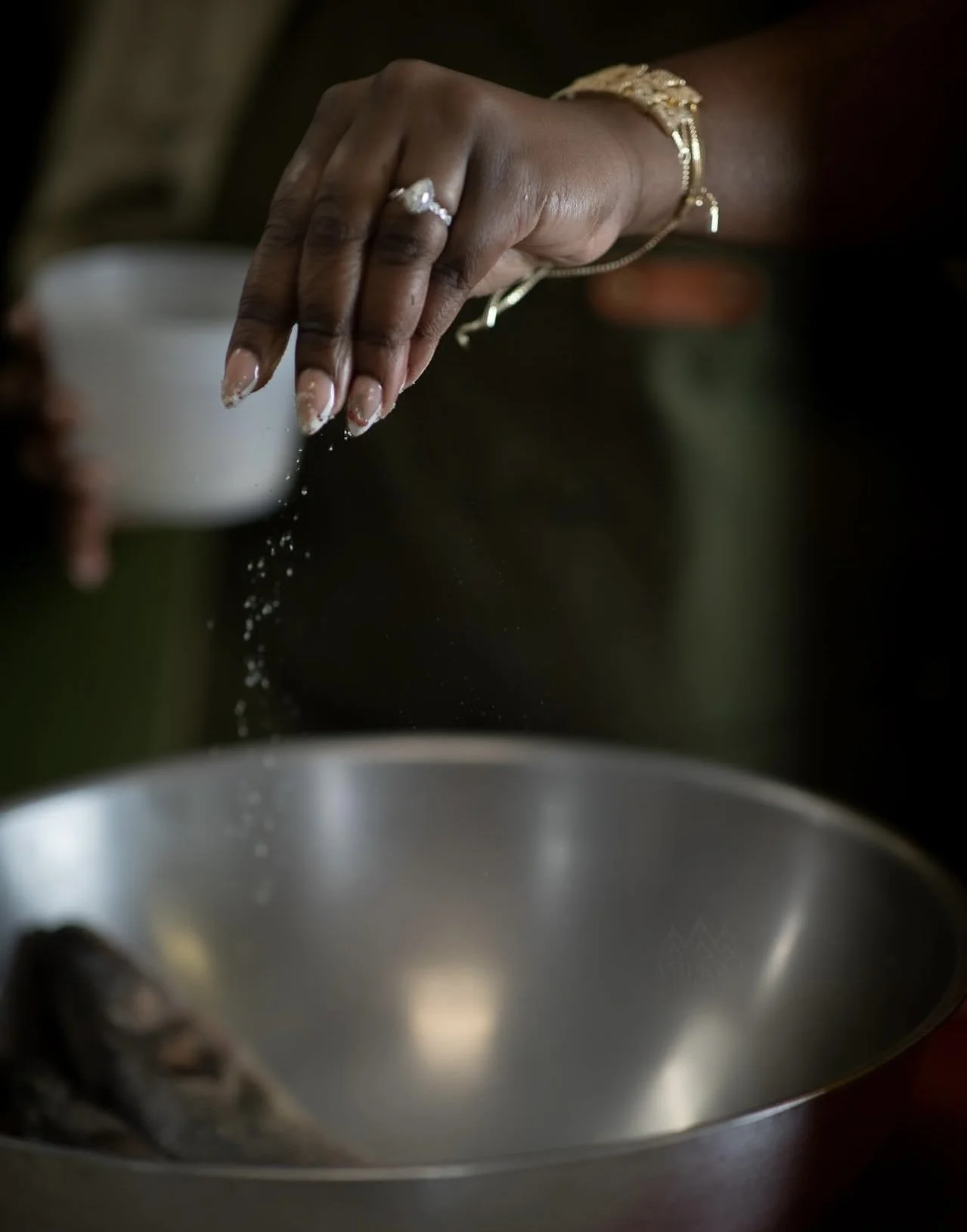 A person sprinkling salt into a metal bowl, wearing rings and bracelets, with salt falling from their hand.