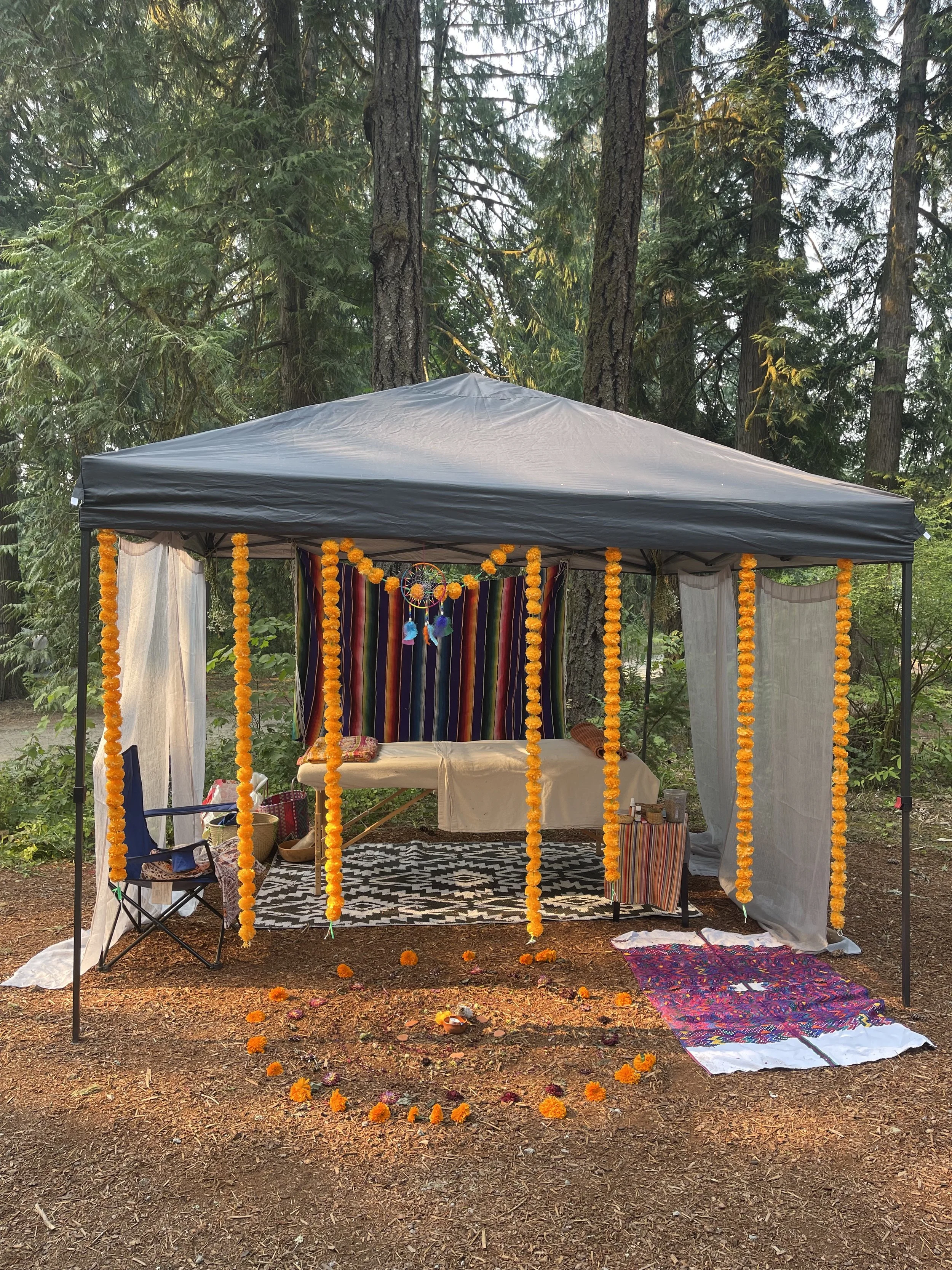 A decorated outdoor canopy with marigold garlands, a hammock, and colorful textiles set up in a forest area with trees and foliage.