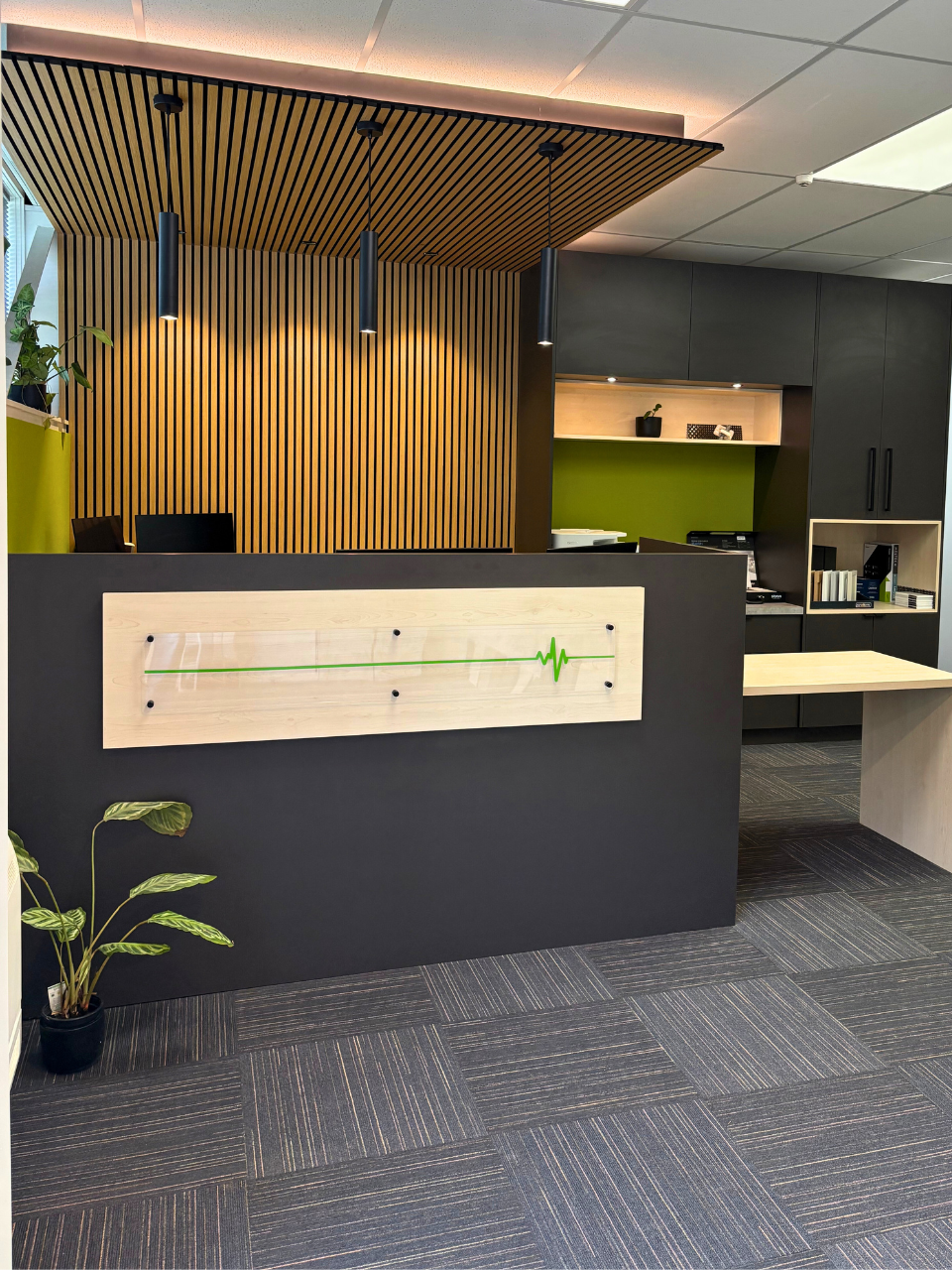 Modern office reception area with black and wood accent wall, green and white wall decor, potted plant, and a digital medical heartbeat display.