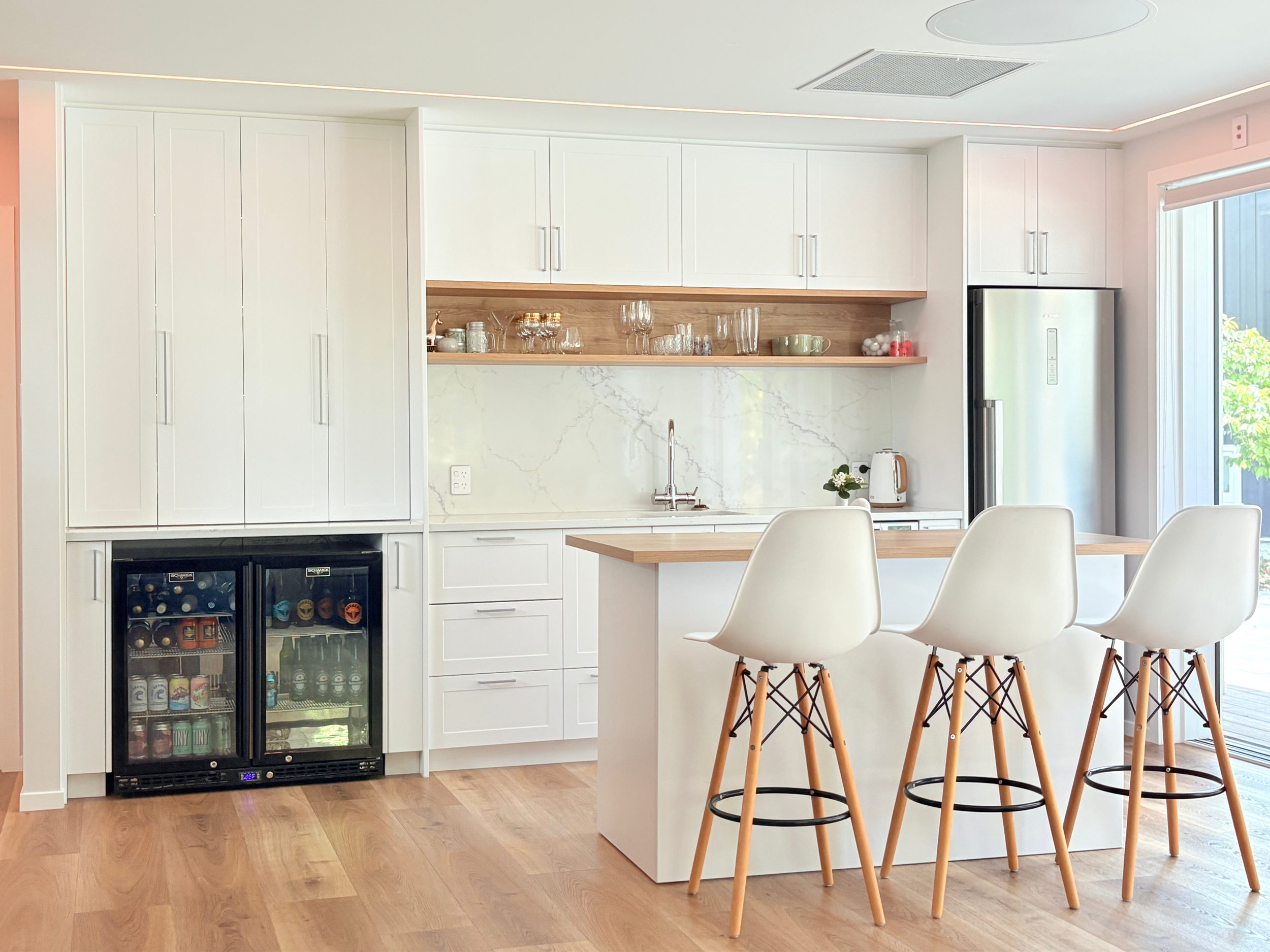 Modern white kitchen with a marble backsplash, open shelves with glassware, a small kitchen island with three white chairs, and a sliding glass door leading outside.