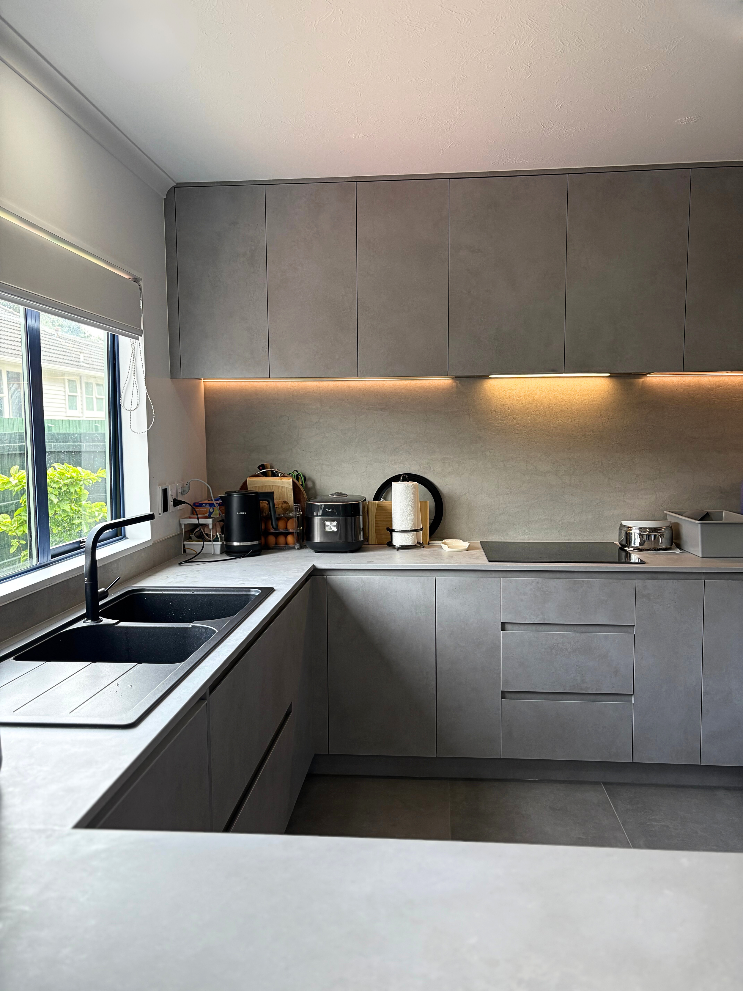 Modern kitchen with grey cabinets, a window with a white blind, black sink, and various small appliances on the countertop.