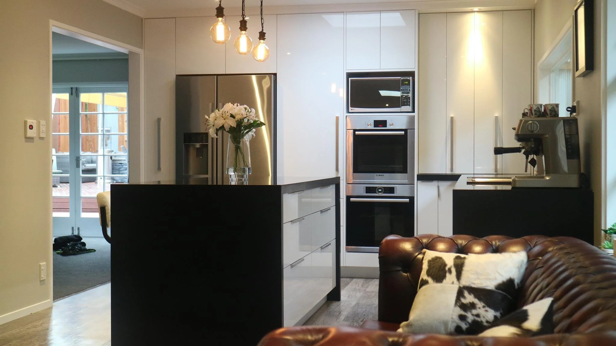 Modern kitchen with white cabinets, stainless steel refrigerator, built-in oven, microwave, coffee machine, and a black island with a vase of white flowers. Part of a leather sofa with cowhide pillows is visible in the foreground.