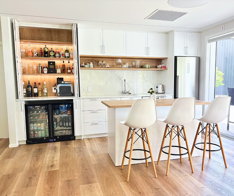 Modern kitchen with white cabinets, wooden accents, marble backsplash, and a breakfast bar with three white chairs. There is a minibar and coffee station on the left, and a refrigerator on the right, with a sliding glass door leading outside.