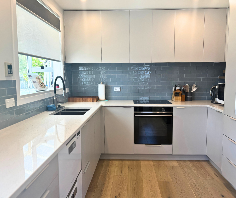 Modern kitchen with white cabinets, gray tiled backsplash, white countertops, a stainless steel oven, and various appliances including a kettle and toaster. There is a window with a blind, a sink with a black faucet, and wooden flooring.