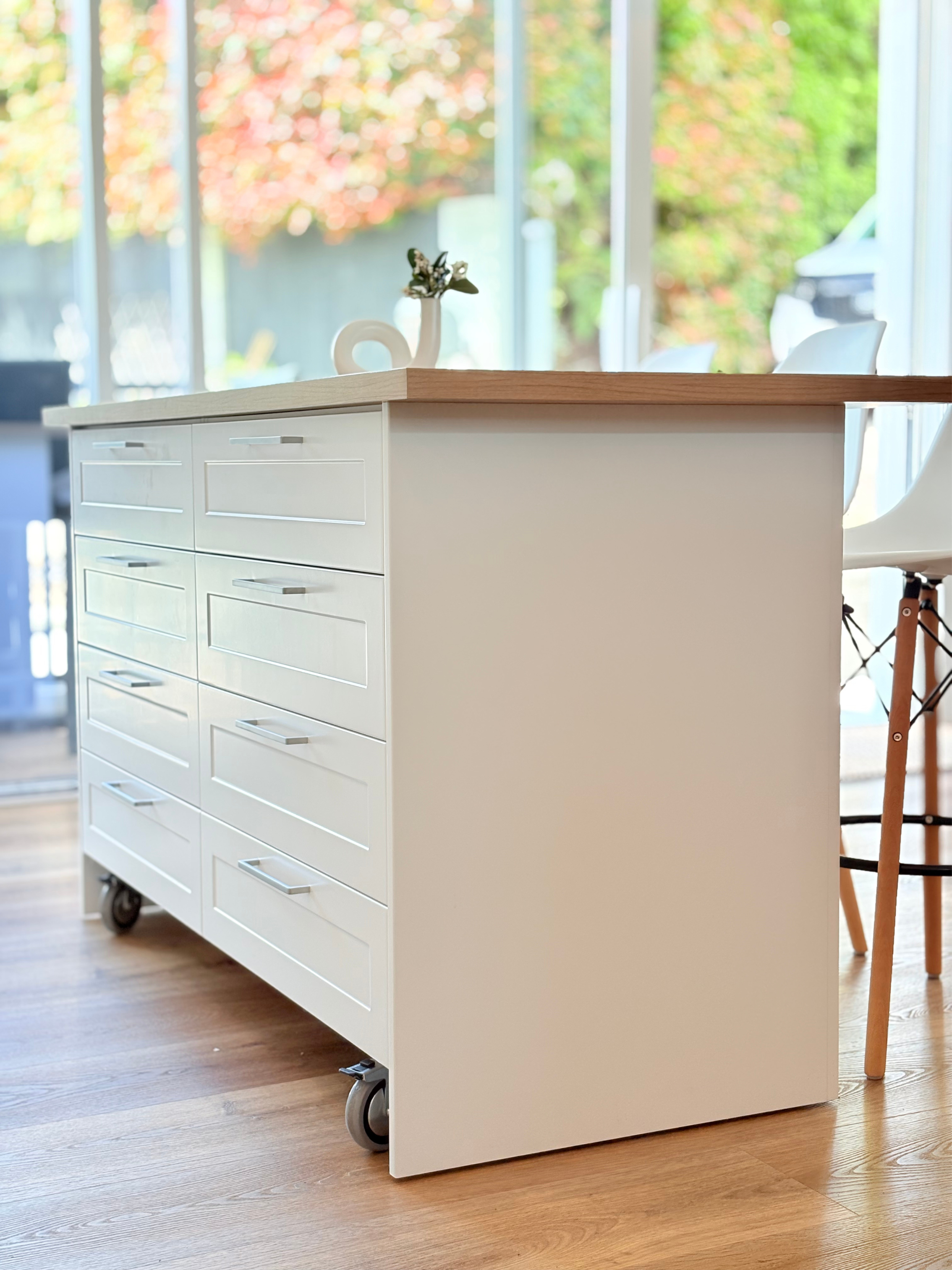 A white kitchen island with a wooden top, decorated with a small white porcelain swan holding a tiny plant, situated in a bright room with large windows and a view of greenery outside.