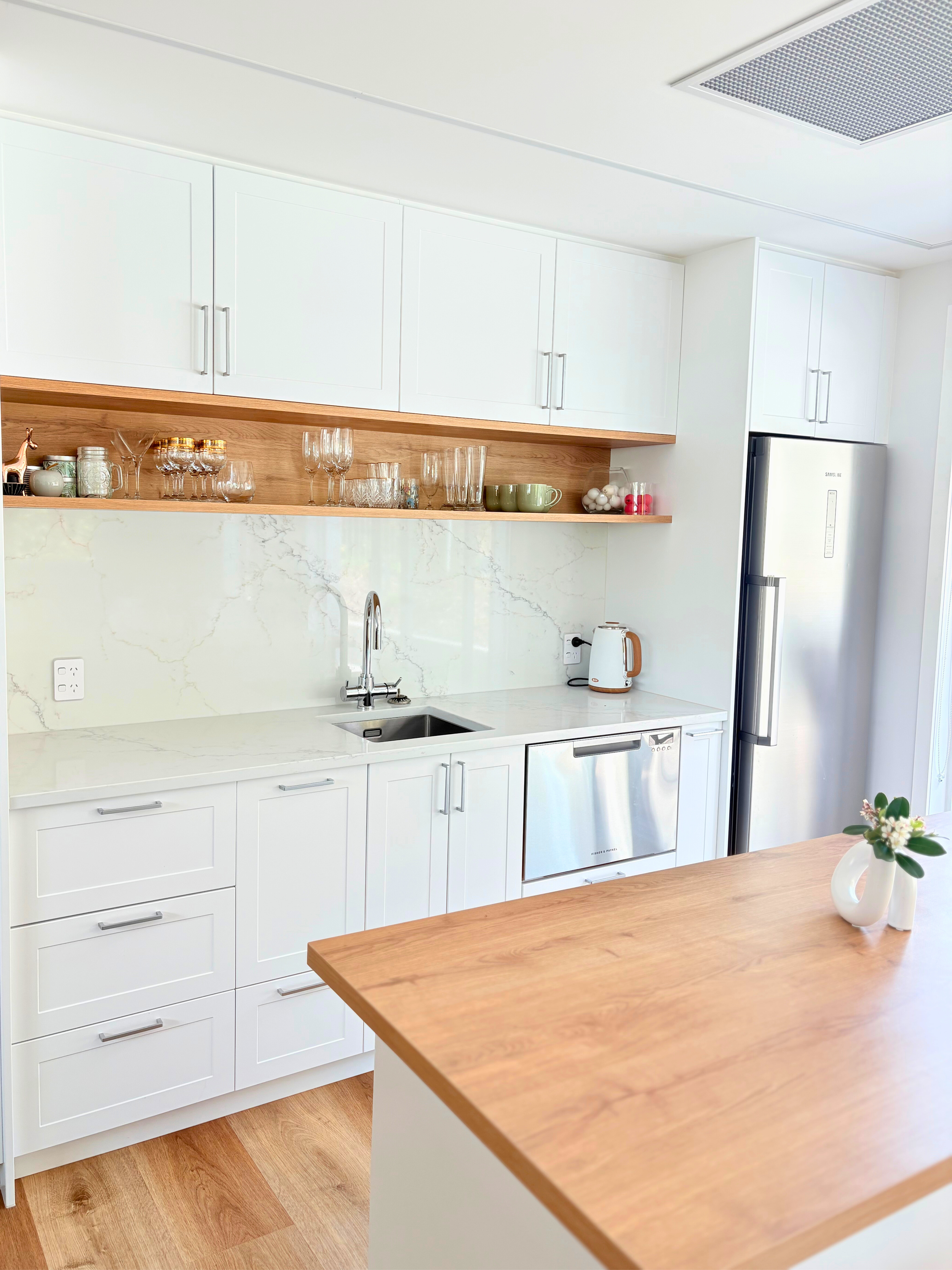Modern kitchen with white cabinets, marble backsplash, wooden open shelf with glassware and bowls, stainless steel refrigerator, counter with a white kettle, and a wooden table with a vase of flowers.