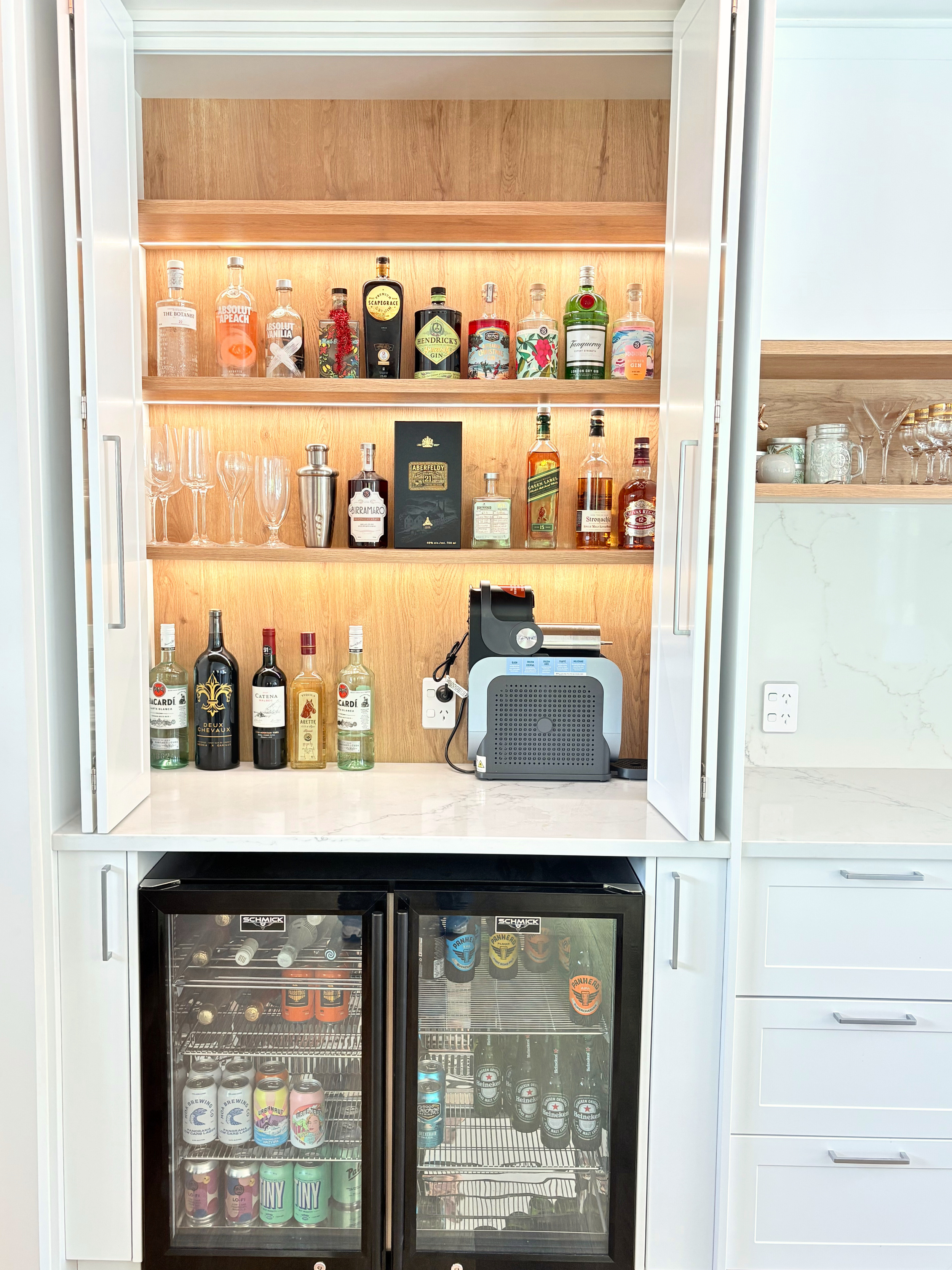 A home bar setup with open cabinets displaying various liquor bottles, glasses, and bar accessories. Below the counter, there is a mini fridge stocked with beverages. A coffee machine is on the counter, and the area is well-lit with under-shelf light