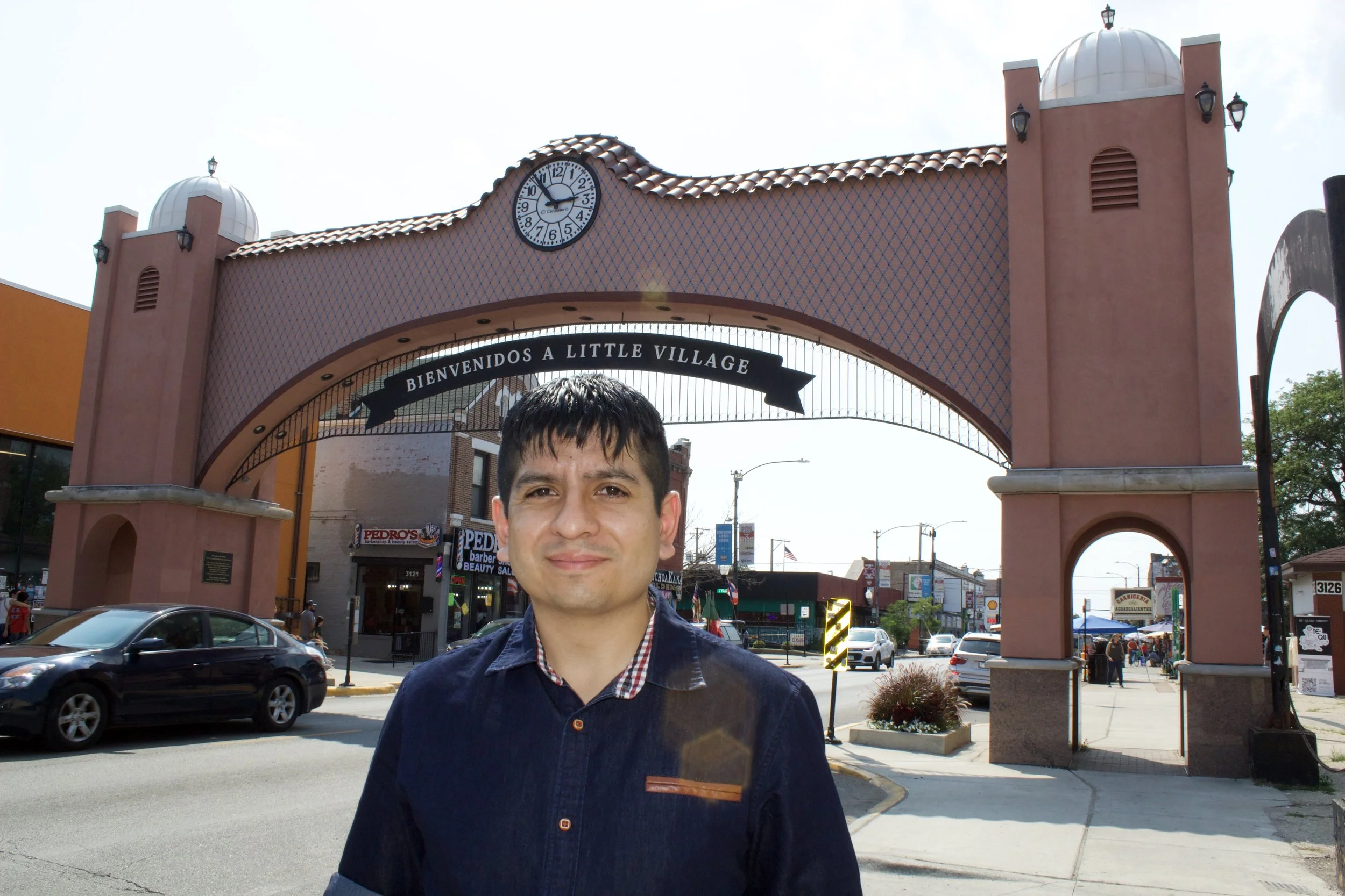 A man standing in front of an archway with a sign that reads "Welcome to Little Village," a clock showing 12:14, and buildings in the background.