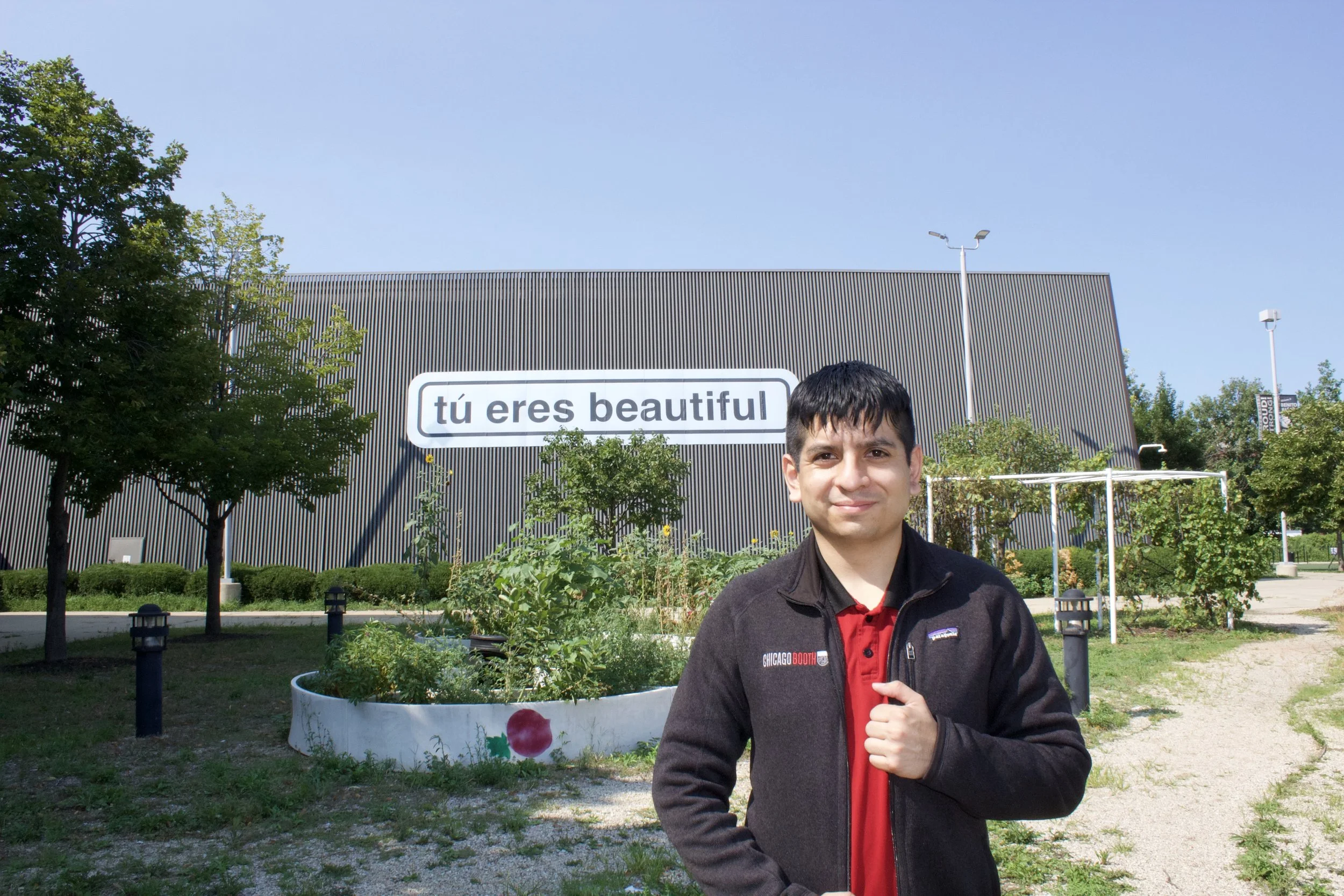 A smiling man standing outdoors in front of a modern building with a sign that reads 'tú eres beautiful.' The man is wearing a black jacket and a red shirt, and there are trees and a landscaped area around him.