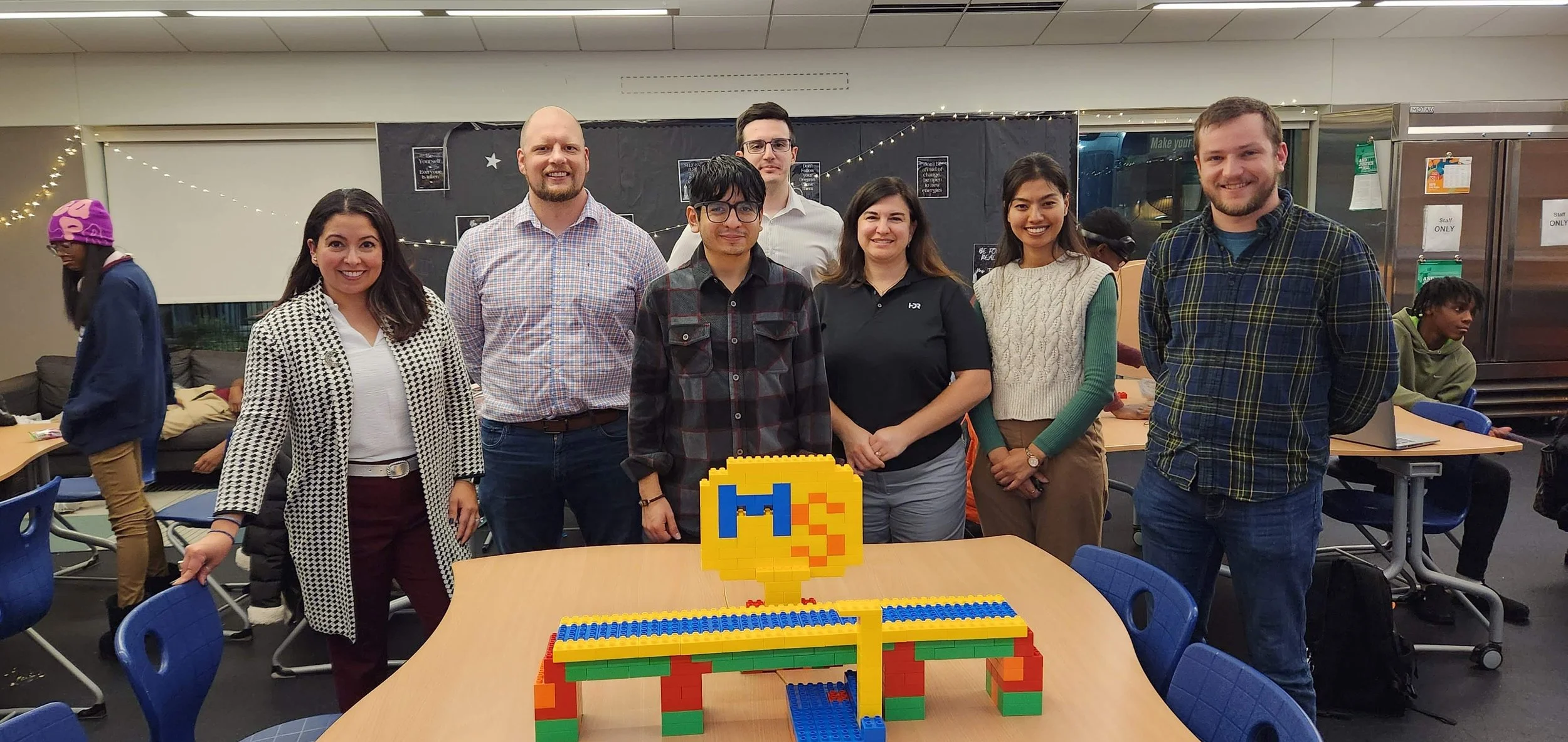 Group of eight people standing behind a table with a LEGO sculpture of a bird on it in a room with black and white decor and string lights.