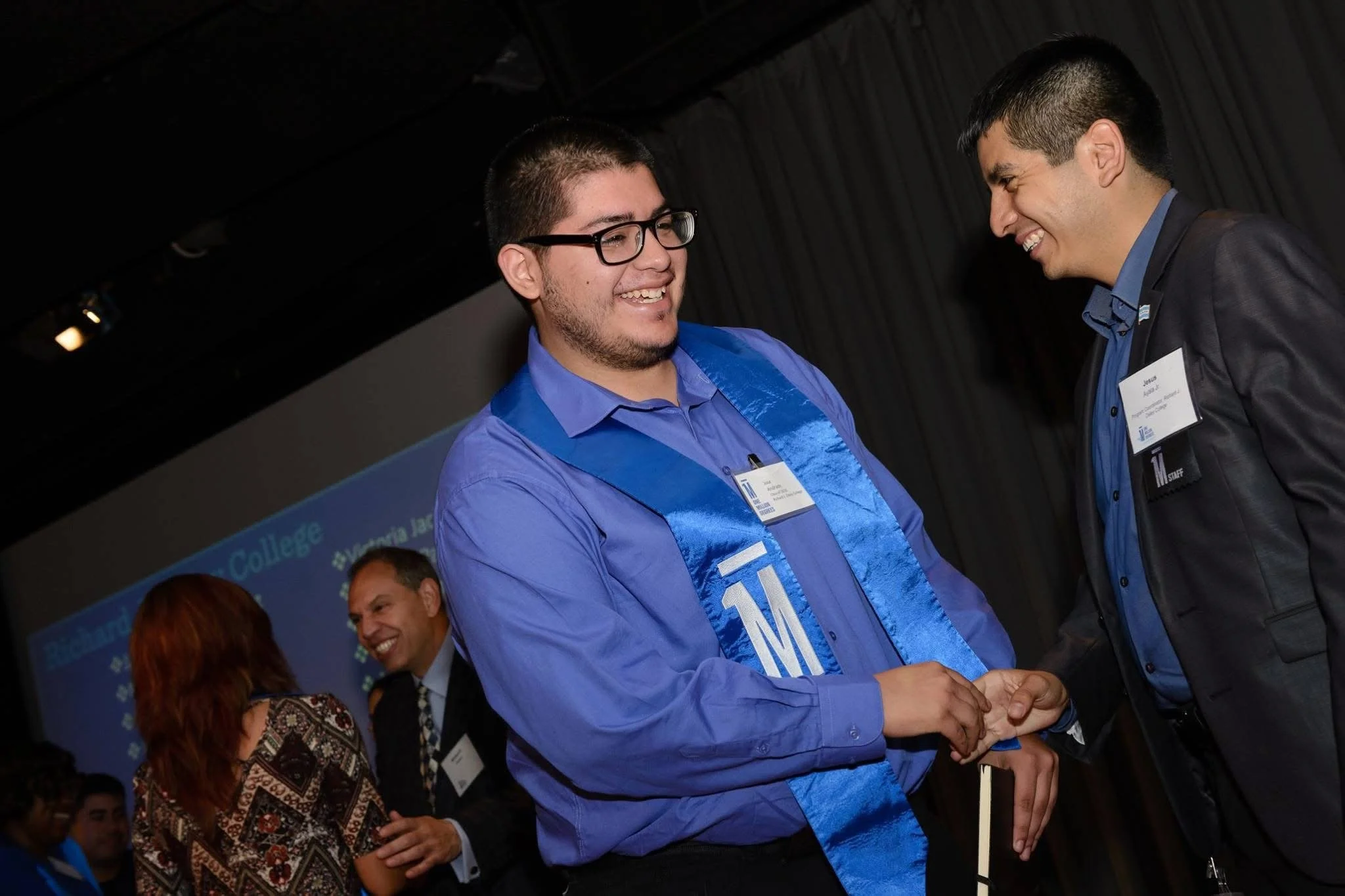 Two men shaking hands at an event, both smiling; one wearing a blue shirt with a sash, the other in dark attire with a name tag.