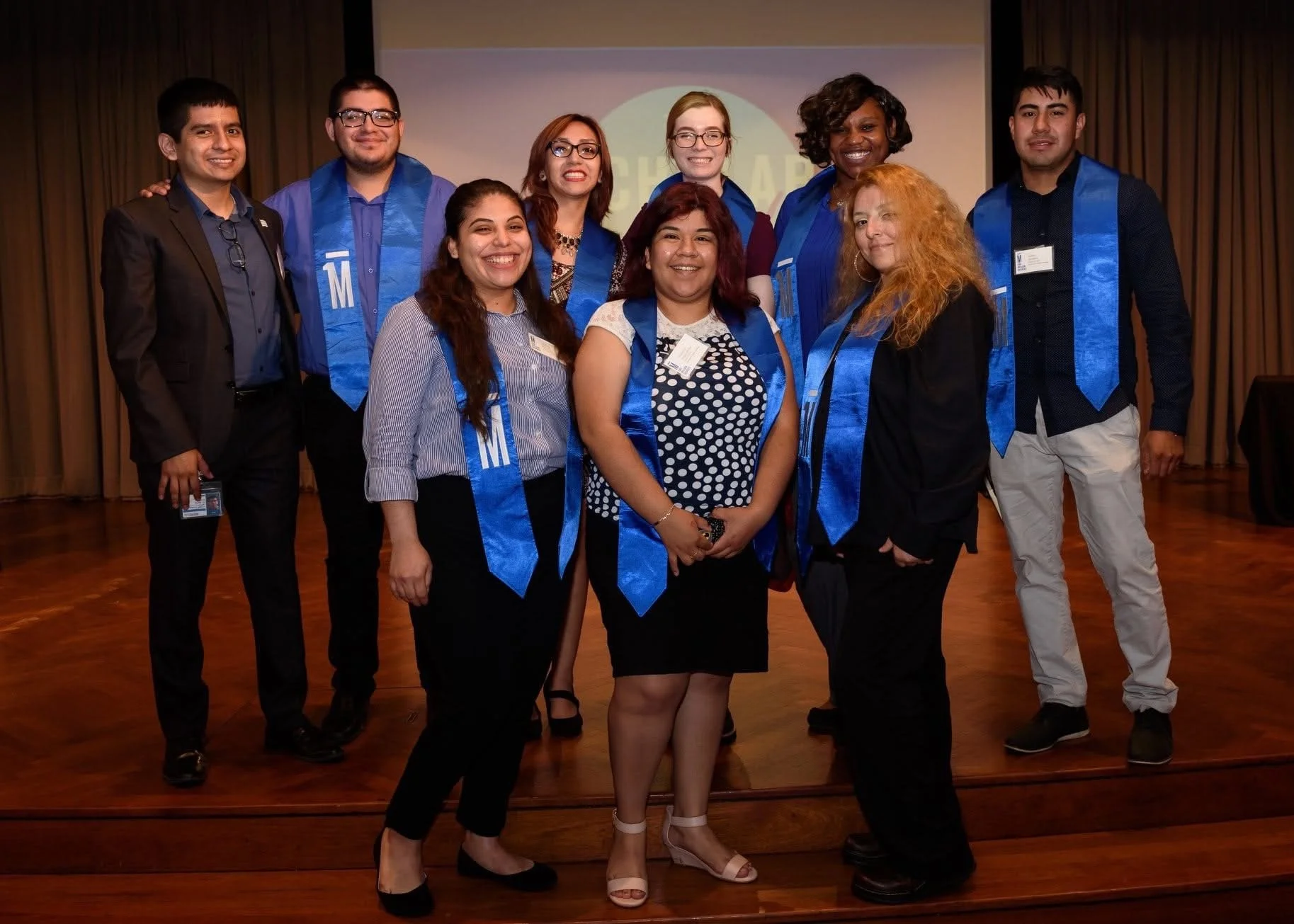Group of nine diverse people standing on a stage, wearing blue sashes and formal attire, smiling at a graduation ceremony or similar event.