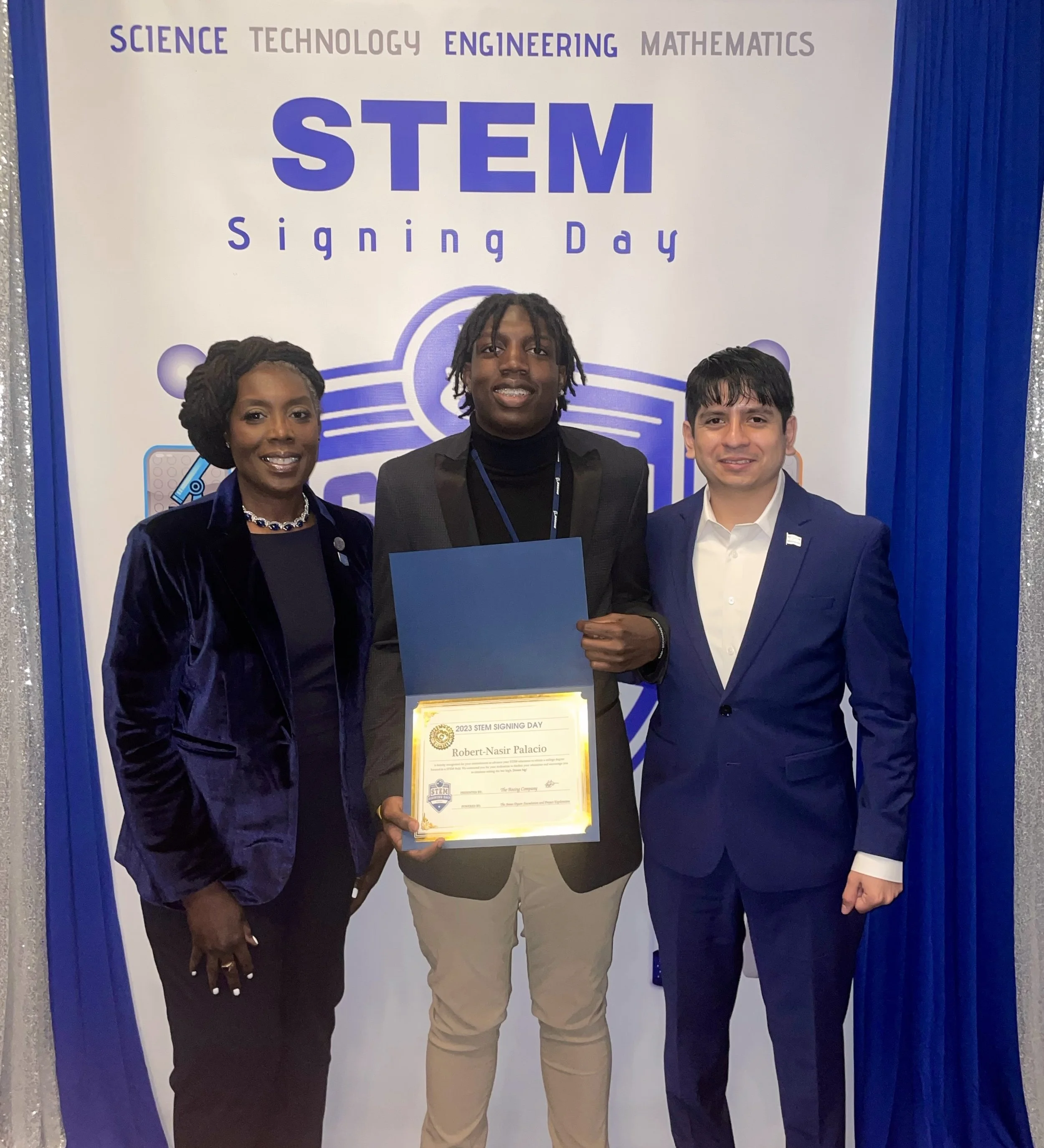 Three individuals standing in front of a blue and white sign that reads 'STEM Signing Day' with the words 'Science,' 'Technology,' 'Engineering,' and 'Mathematics' at the top. One person is holding a certificate.