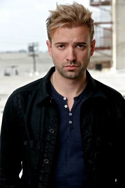 Young man with blond hair and a serious expression standing outdoors in front of a cityscape with buildings and power lines.