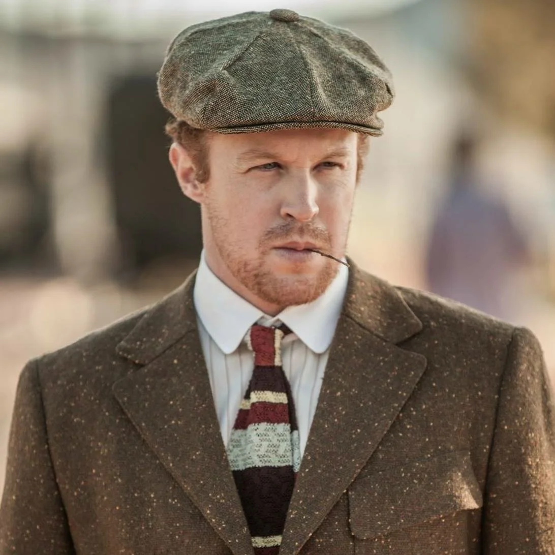 A man with red hair and a beard wearing a brown tweed flat cap, a brown speckled blazer, a white collared shirt, a striped tie, and holding a toothpick in his mouth.
