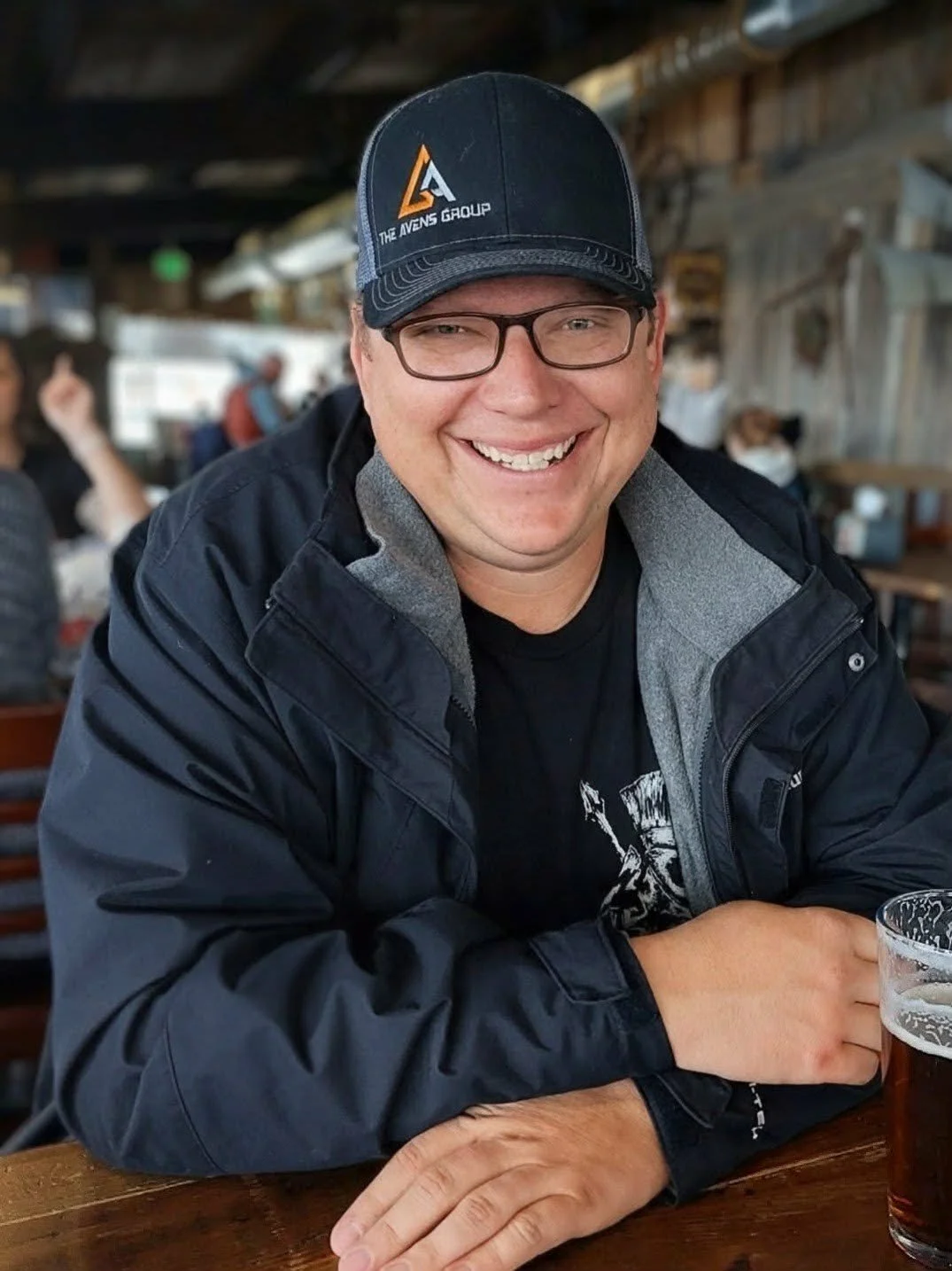 A man wearing glasses and a black cap with a logo, smiling, sitting at a table in a casual restaurant or bar, with a beer in front of him.