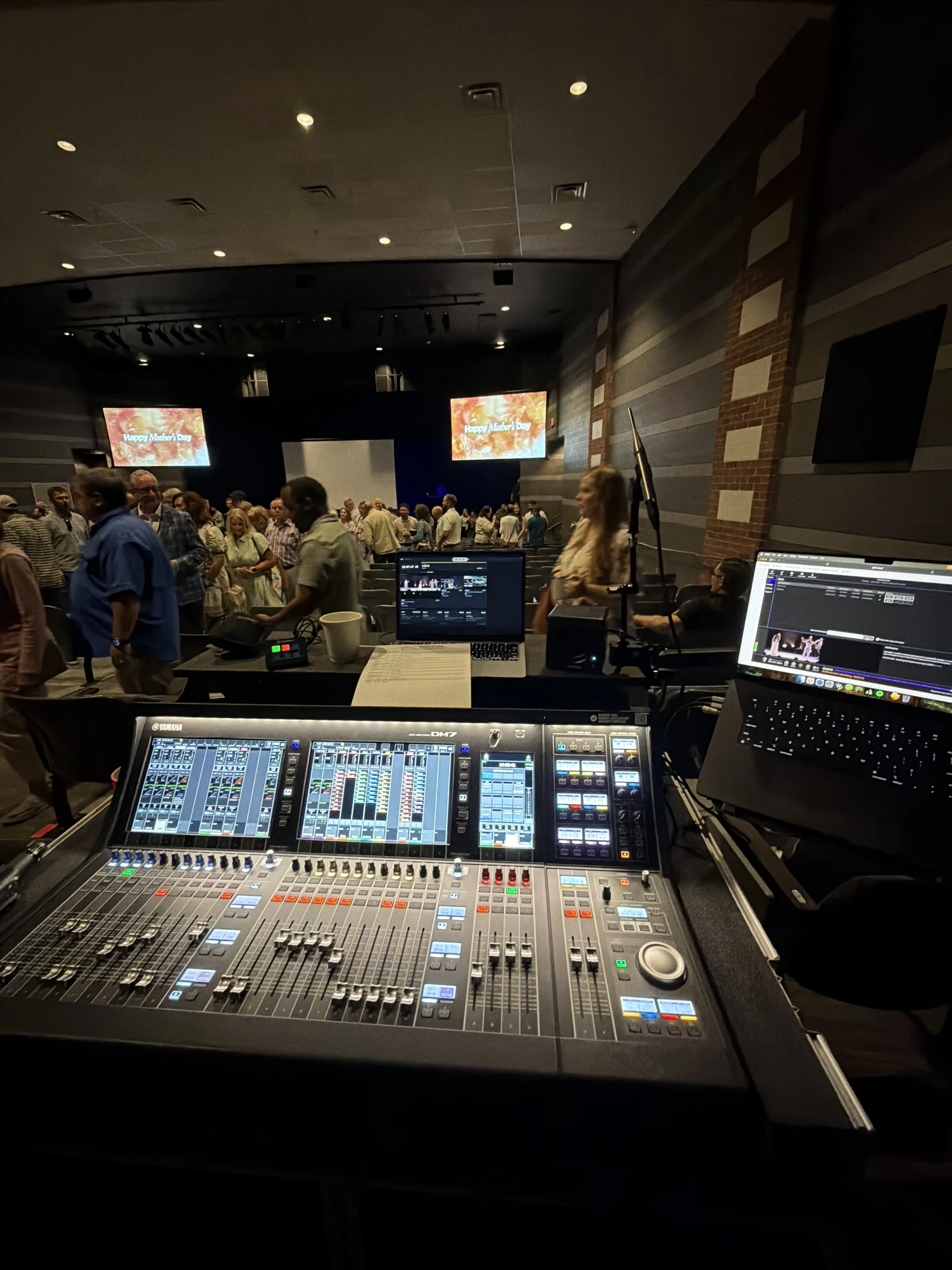 Sound mixing console in the foreground, with a computer screen on the right and a stage with people in the background; two screens display a message "Happy Mother's Day" at an indoor event.