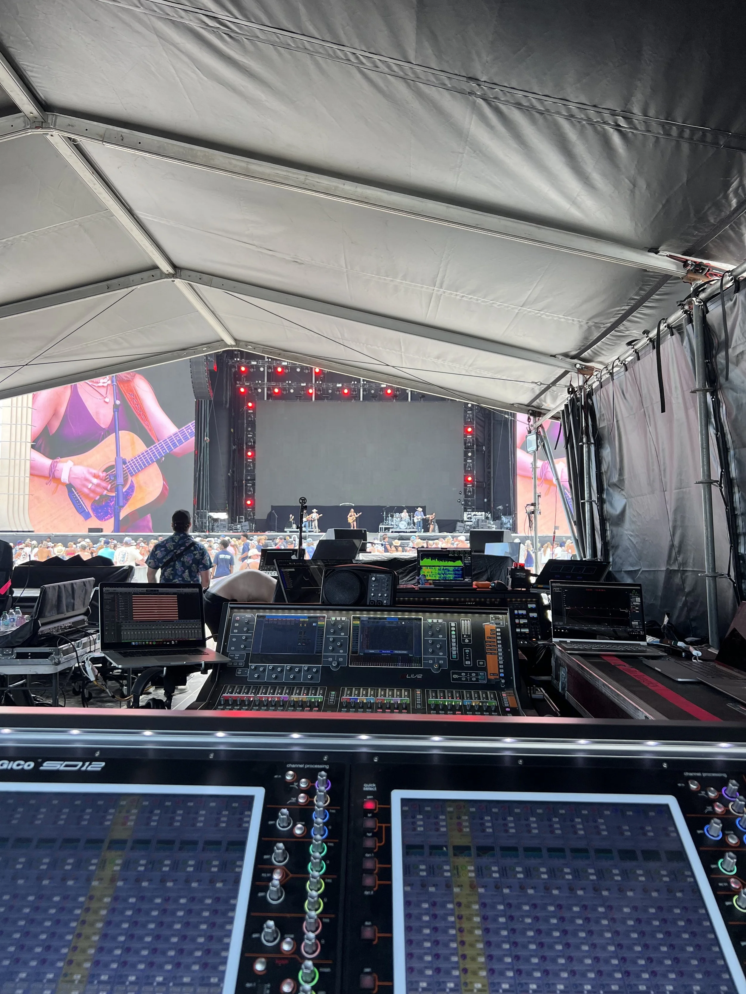 Inside view of a stage at a concert with a sound mixing console, audio equipment, and two large screens showing a person playing guitar and singing.