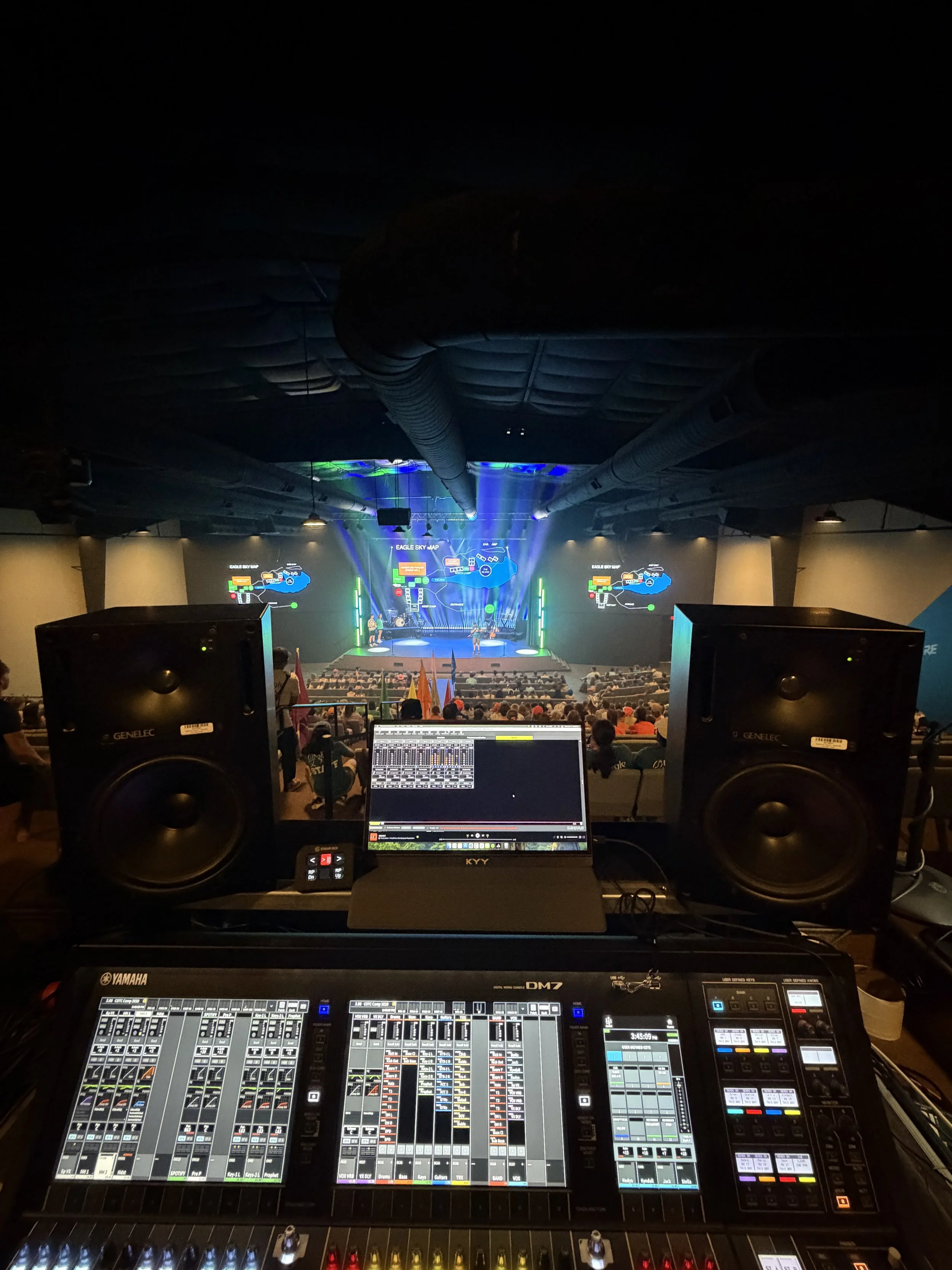 View from a music production booth overlooking a stage with a band performing illuminated with colorful lights, audience in seats, and large screens displaying maps and graphics.