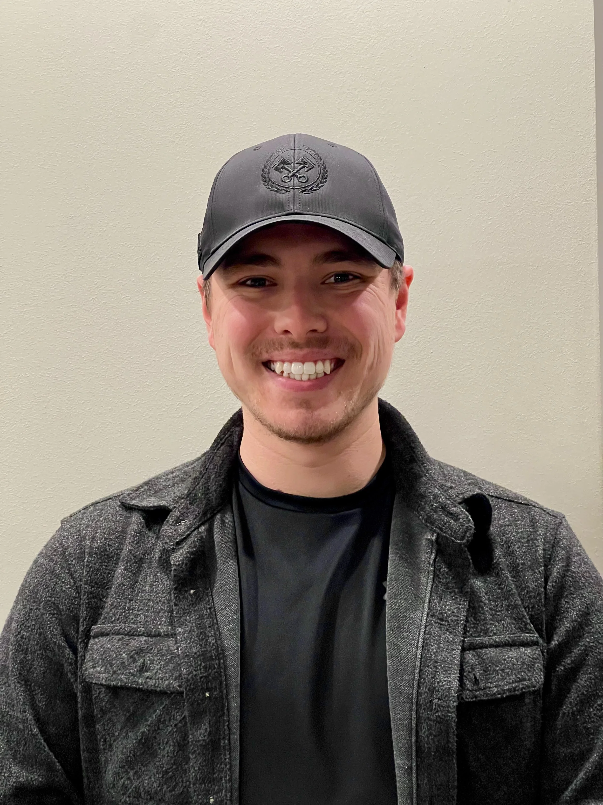 A young man smiling, wearing a dark gray baseball cap with a logo, a dark shirt, and a gray jacket, standing against a plain beige wall.