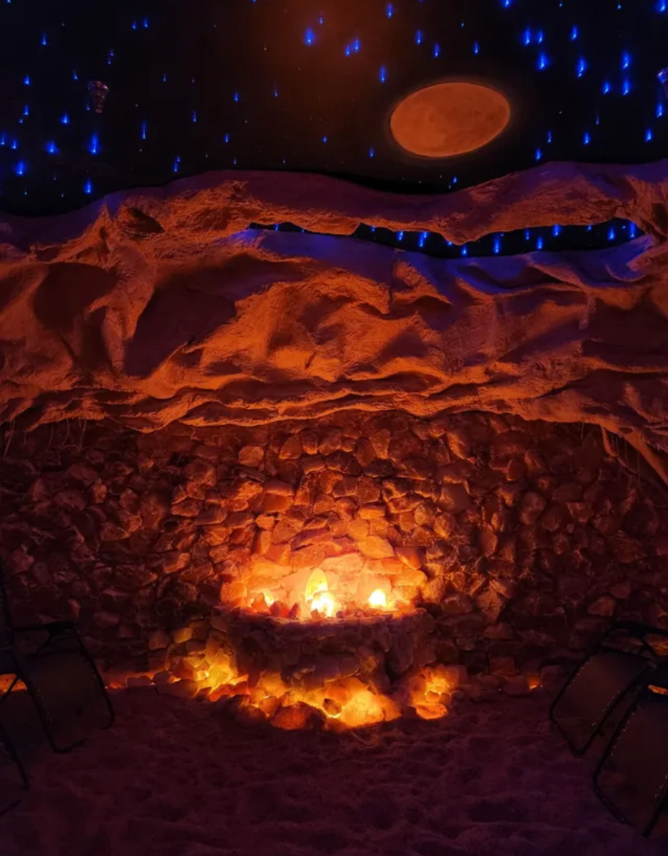 A salt cave with a glowing himalayan salt lamps surrounded by boulders and salt. The ceiling features a starry night sky with twinkling lights and a full moon.