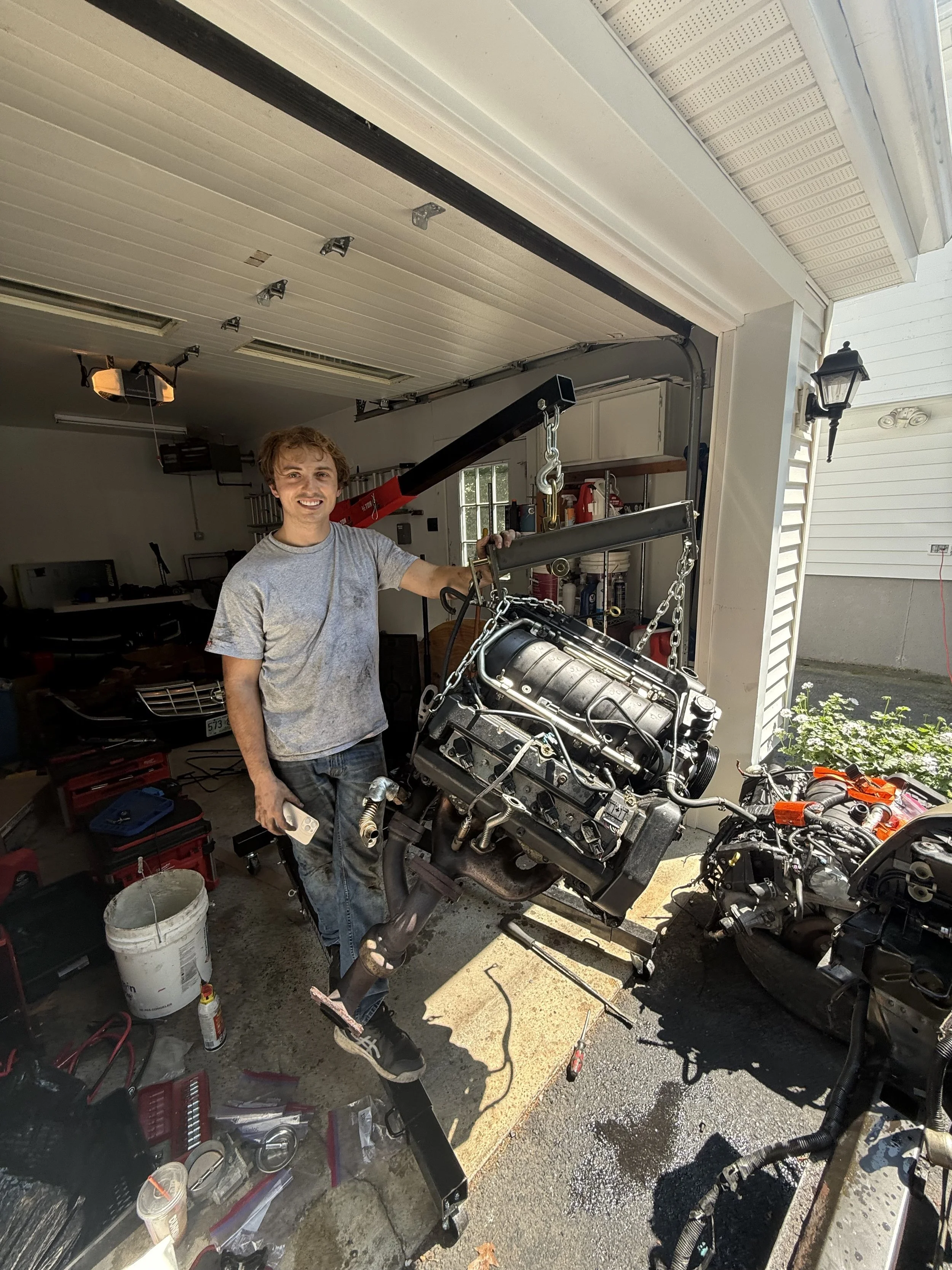A man is working on a motorcycle engine in a garage. He is smiling, holding a tool, with motorcycle parts around him and a water bucket and various tools on the ground.