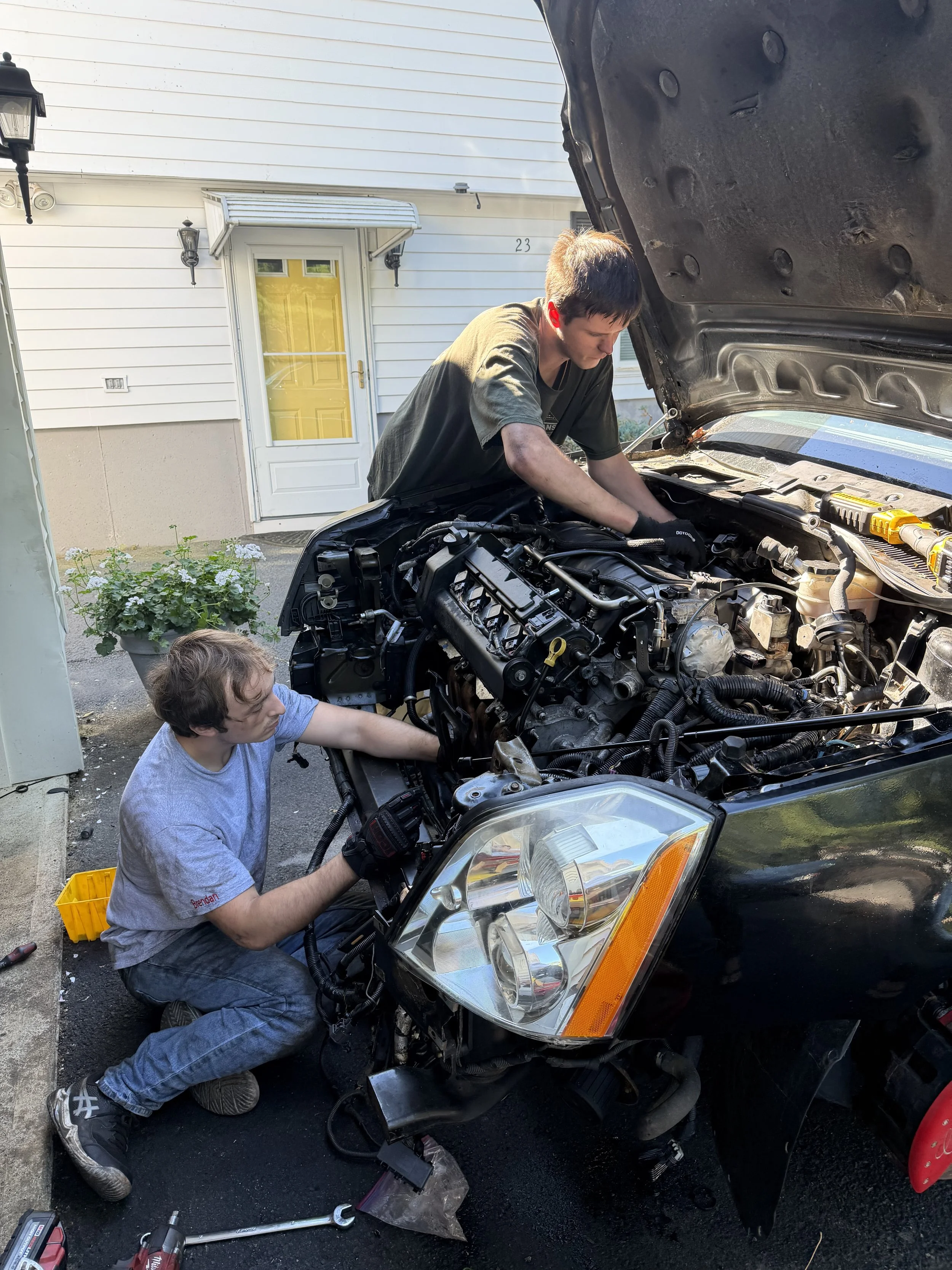 Two young men repairing a car with the hood open outside a white house with yellow door. One is crouching by the front wheel, the other is leaning over the engine. Tools are on the ground.