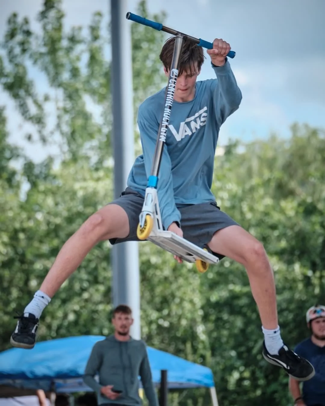 A young man in a blue Vans sweatshirt and shorts on a scooter jumps mid-air, with his legs spread wide, during an outdoor event. Two other individuals and trees are visible in the background.
