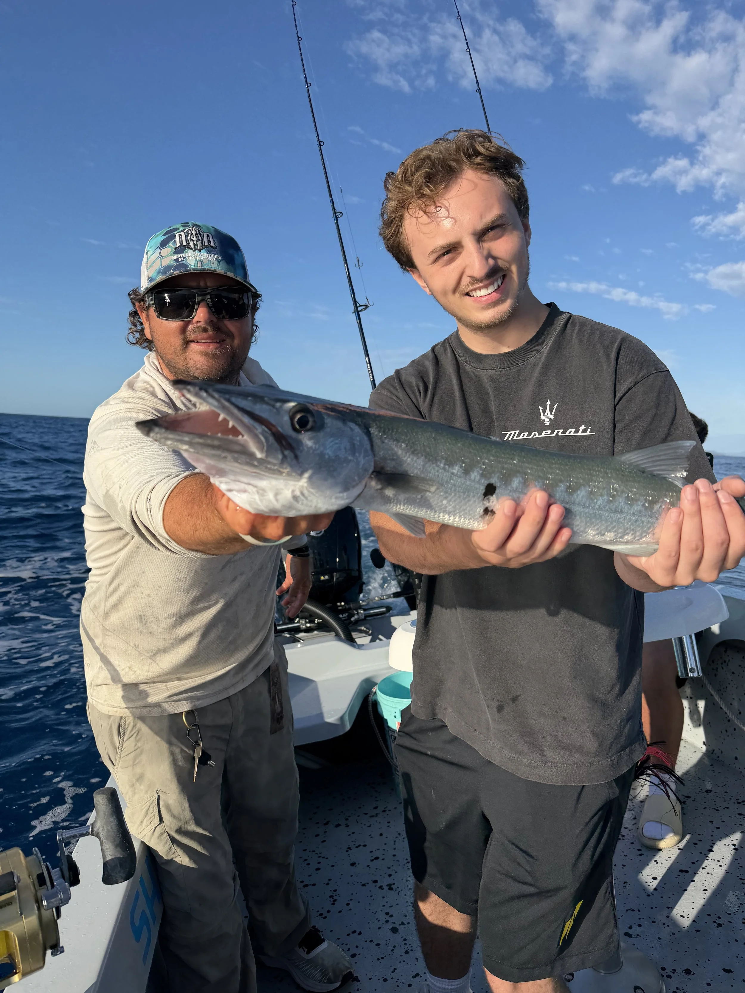 Two men on a boat holding a large fish, smiling at the camera with fishing rods in the background and blue sky overhead.