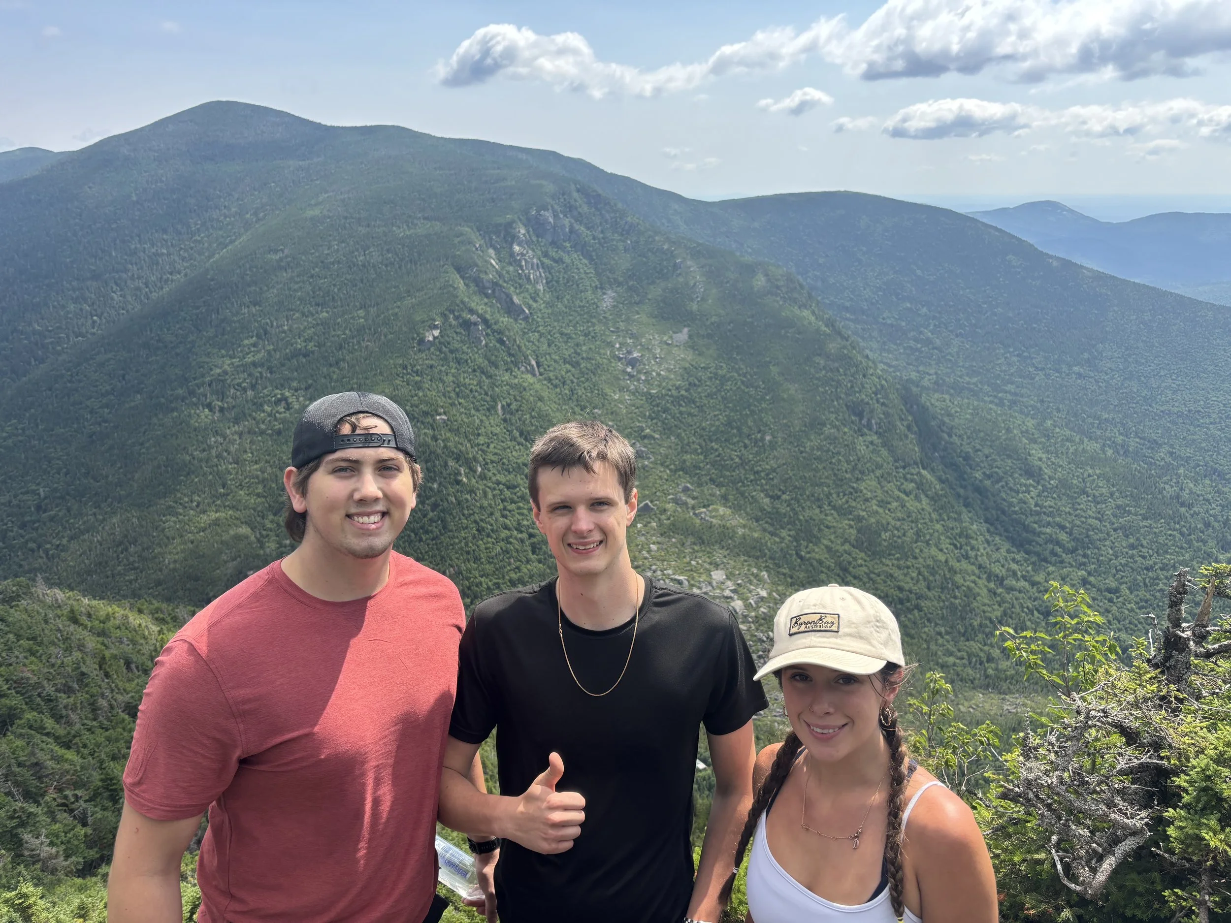 Four young people smiling in front of mountains and green landscape during daytime, one showing a thumbs-up.