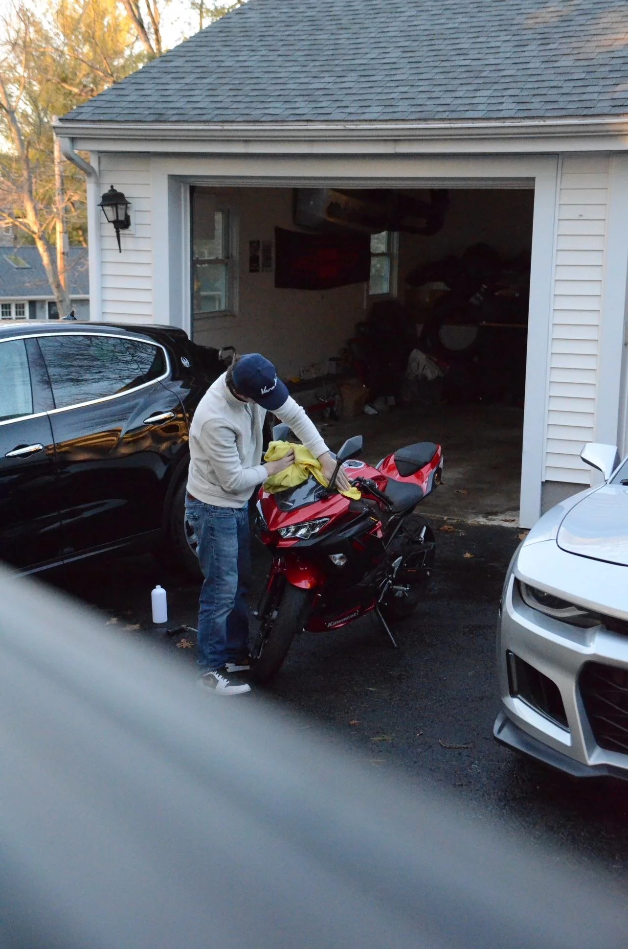 A person wearing a white hoodie and cap cleaning a red motorcycle in a driveway, with a black car parked beside him. The garage door is open, revealing stored items inside. The scene takes place outdoors in daylight, with trees visible in the background.