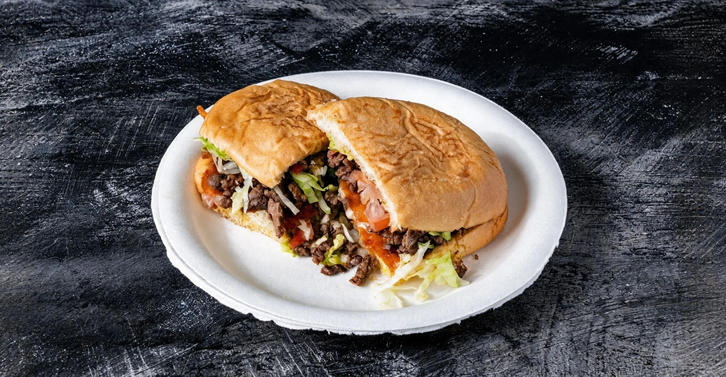 A sandwich with ground beef, lettuce, tomato, and sauce, served on a white foam plate.
