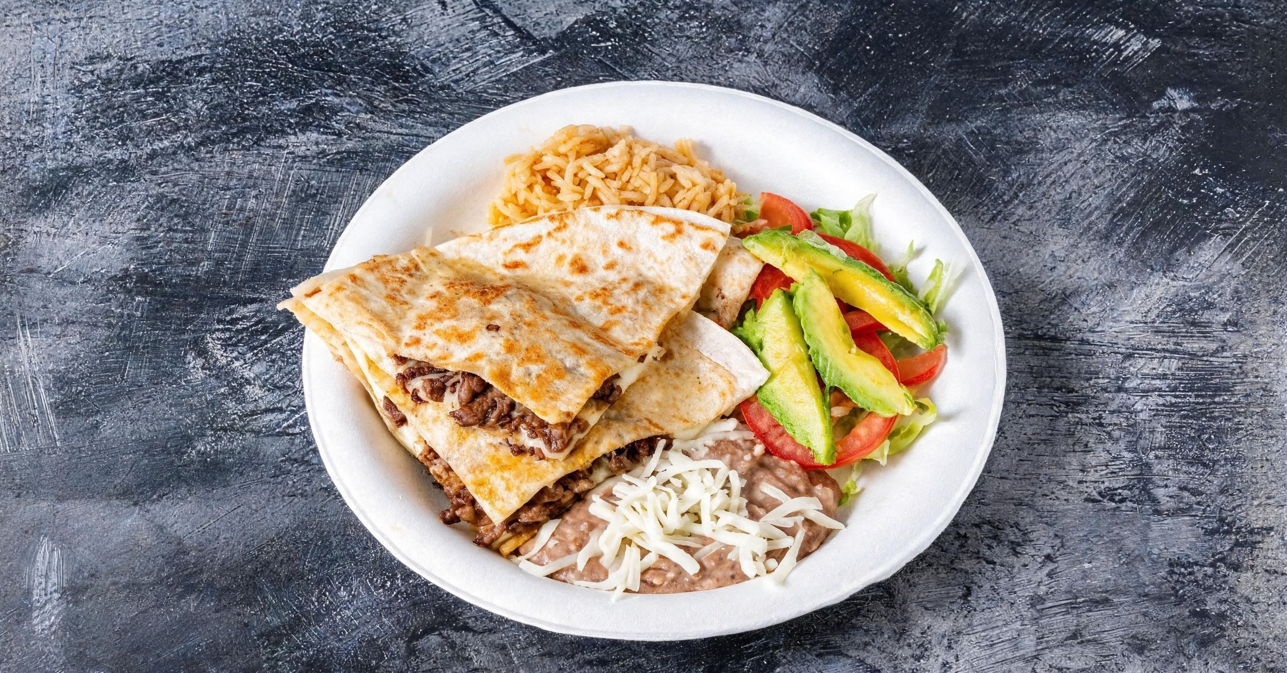 Plate of Mexican food including a beef quesadilla, rice, refried beans with cheese, and a salad with avocado, tomato, and lettuce, on a textured dark surface.