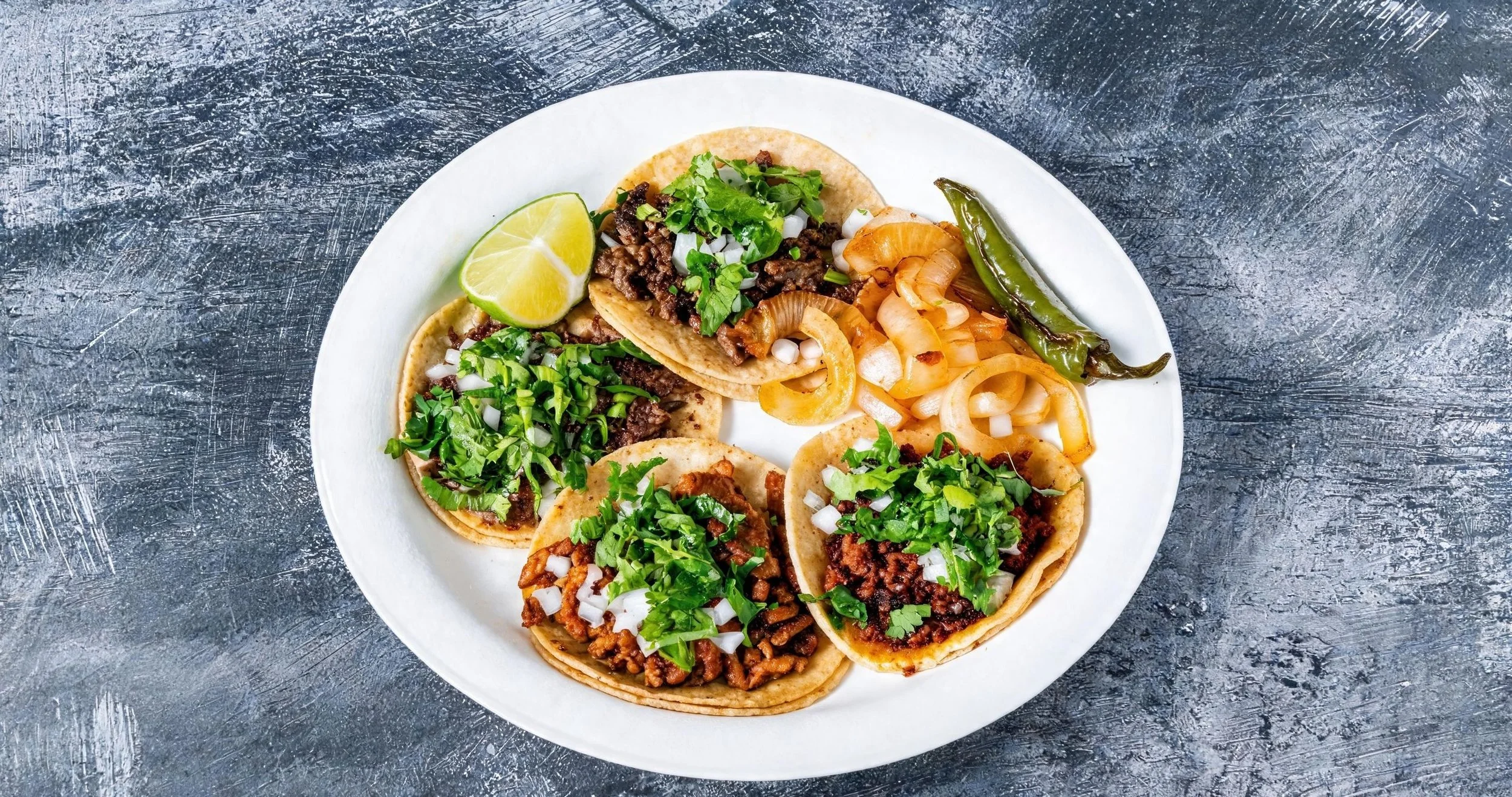 A white plate with five tacos filled with seasoned ground meat, chopped onions, cilantro, and shredded cheese, with a lime wedge on the side, on a textured gray surface.