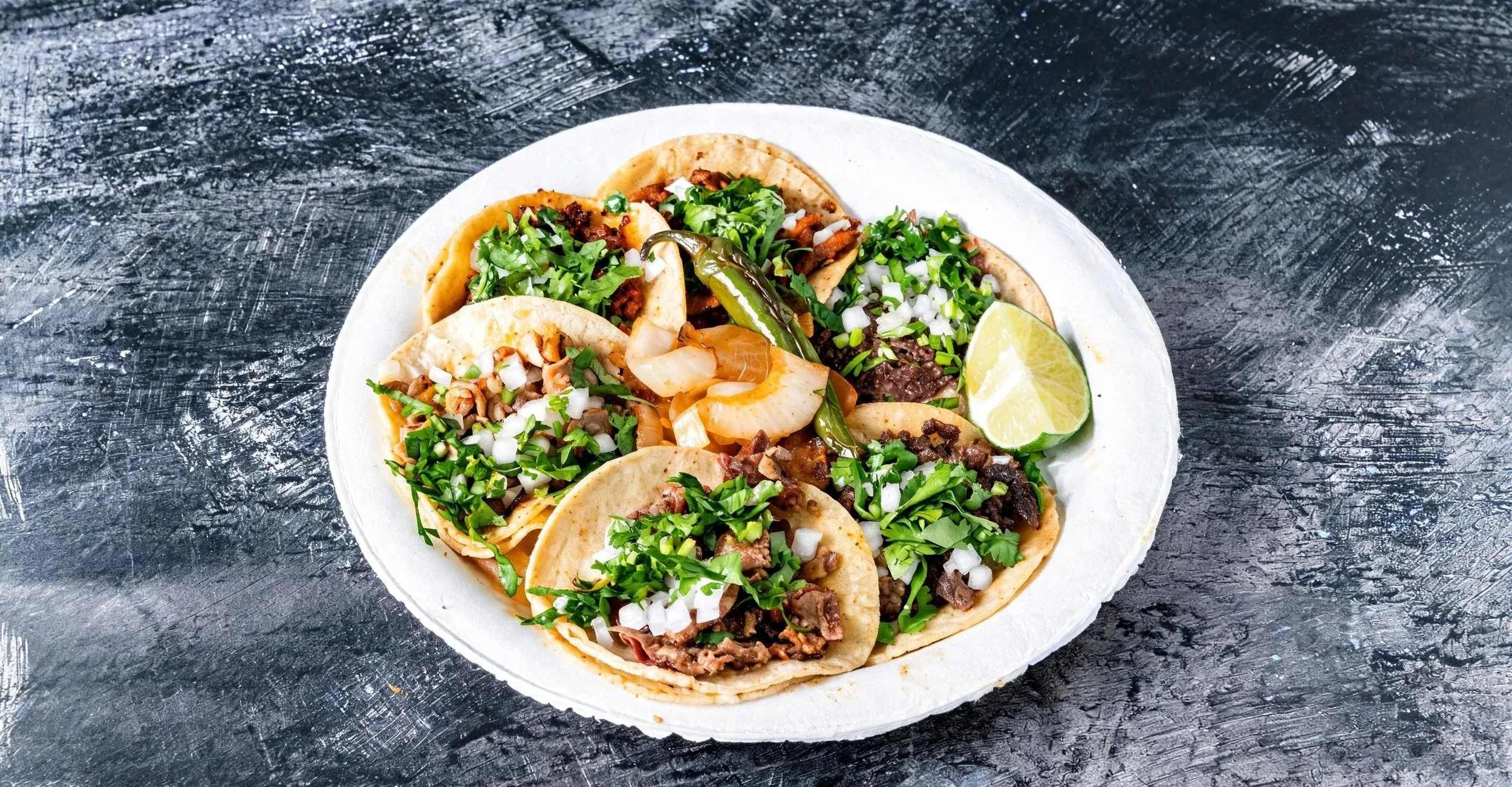 Plate of tacos filled with beef, chopped onions, cilantro, and sliced lime wedge on a dark textured surface.