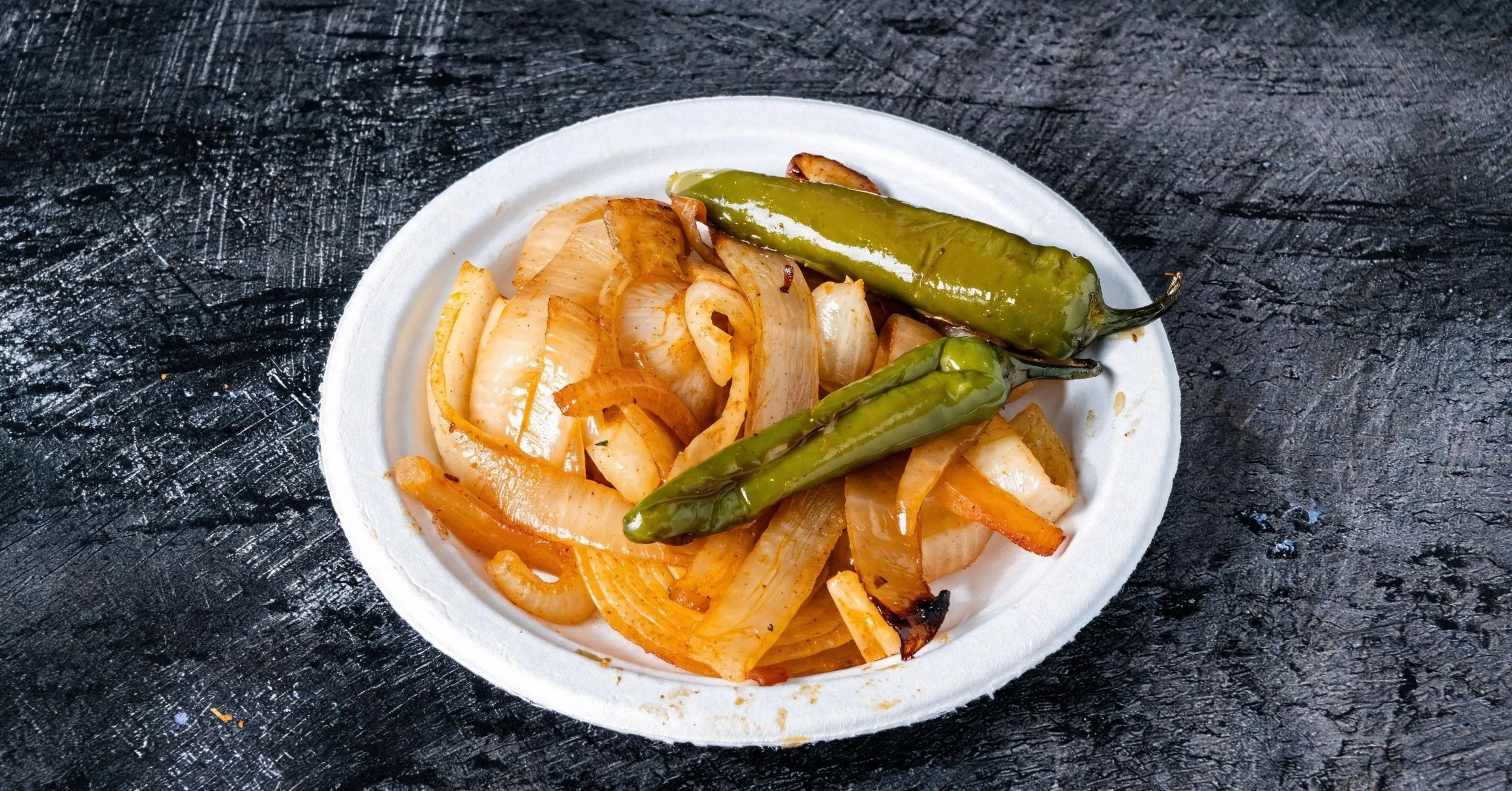 A white disposable plate with grilled onions and two grilled jalapenos on a dark textured surface.