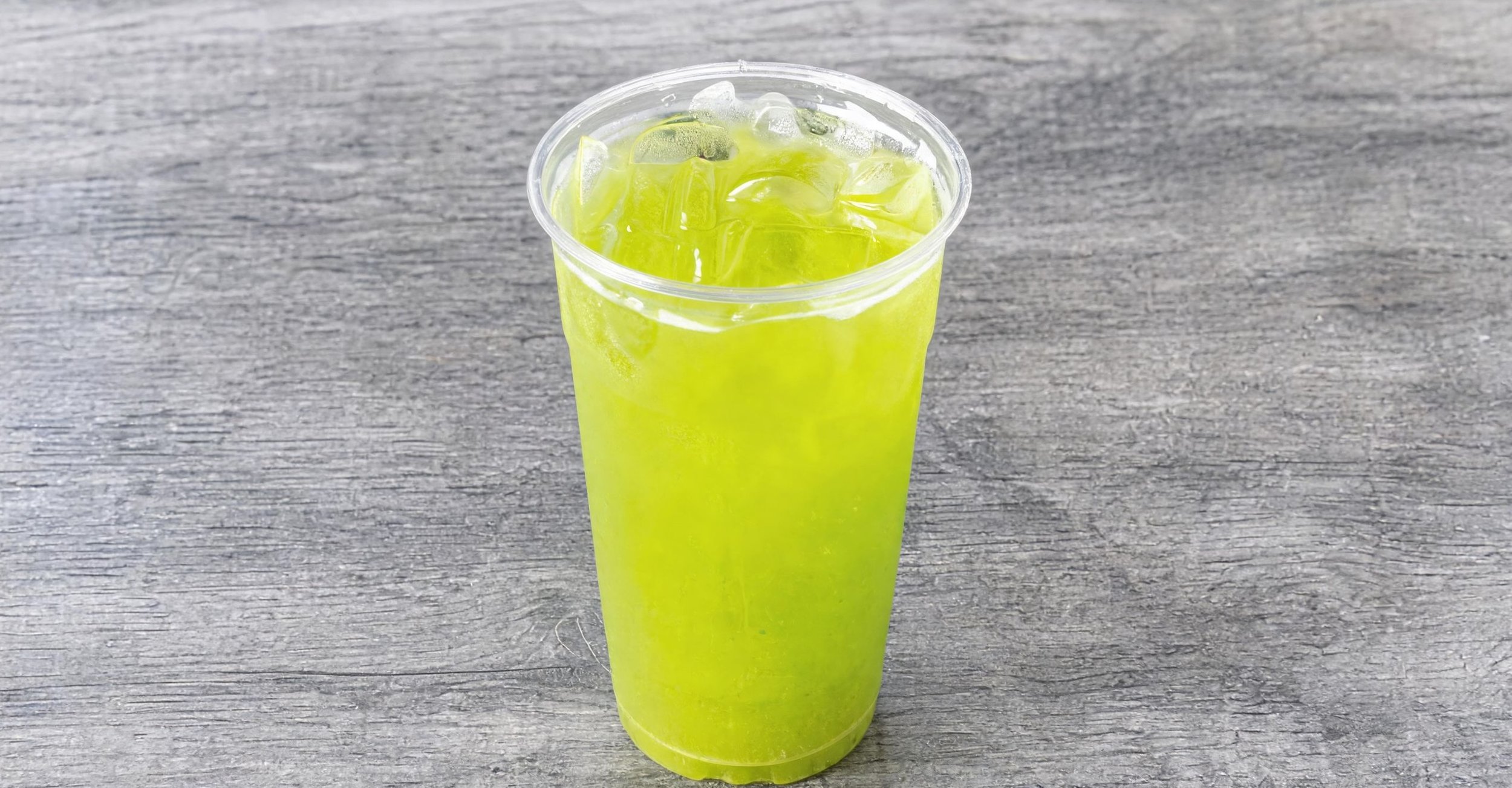A clear plastic cup filled with bright yellow-green beverage and ice cubes, placed on a gray wooden surface.