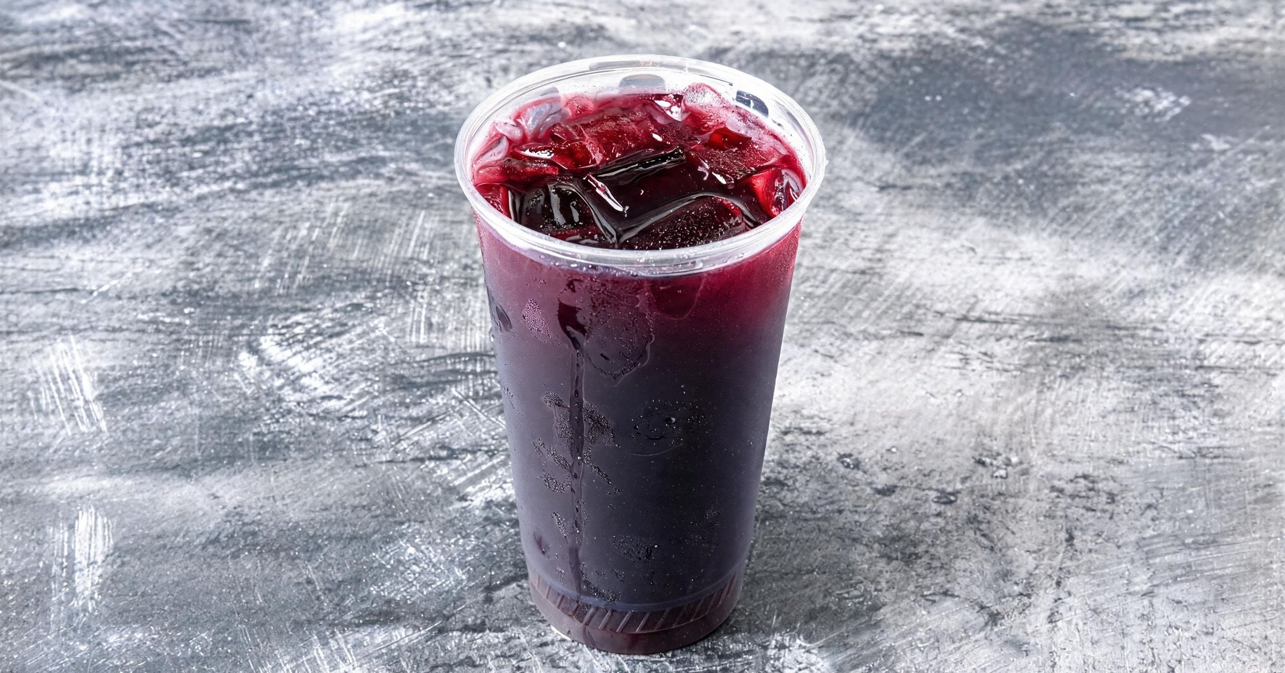 A clear plastic cup filled with dark-colored iced beverage, sitting on a textured gray surface.