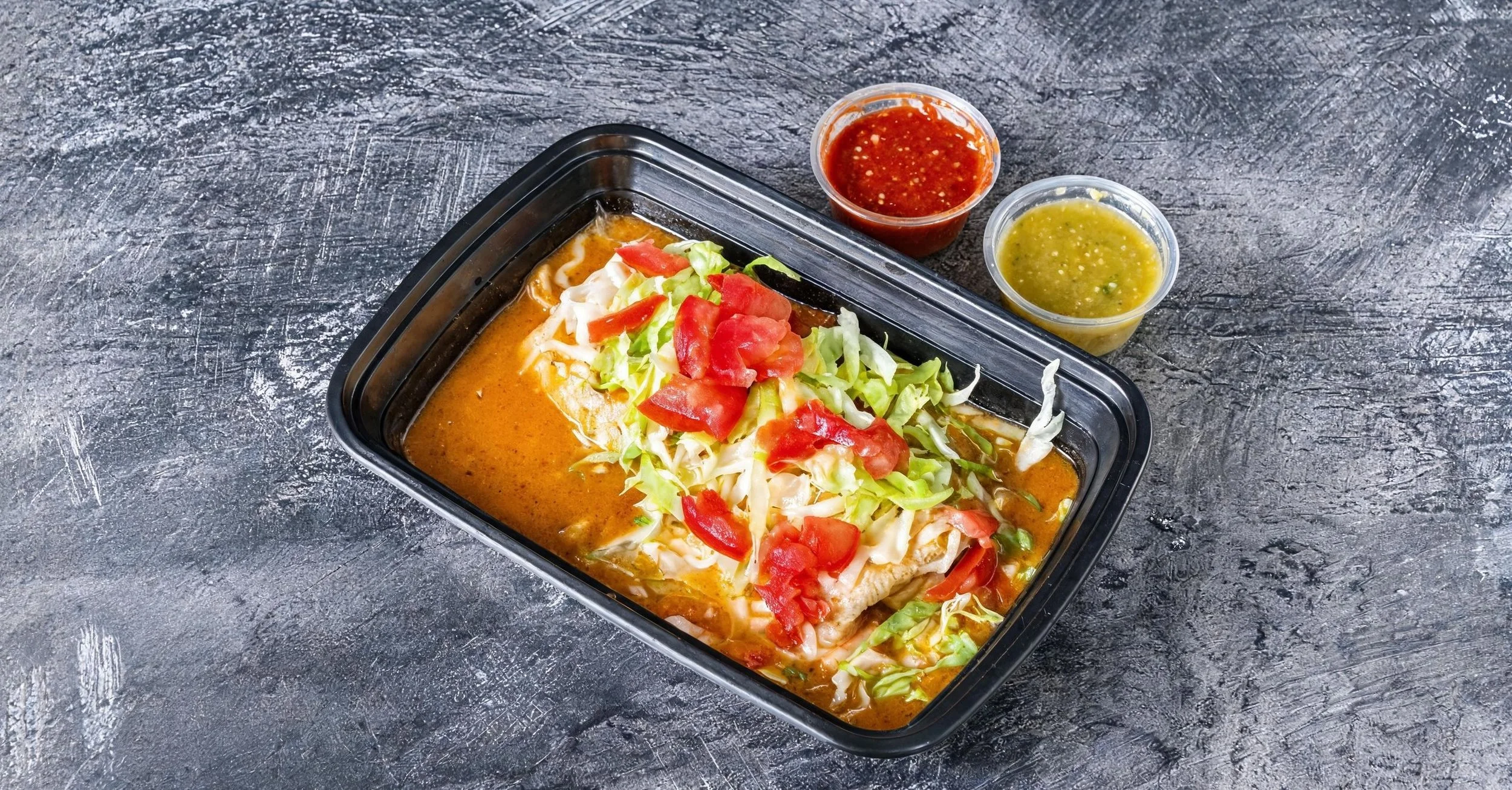 A takeout container filled with Mexican food, topped with shredded lettuce, chopped tomatoes, and sauce, with two small cups of salsa and green chili sauce on a gray textured surface.