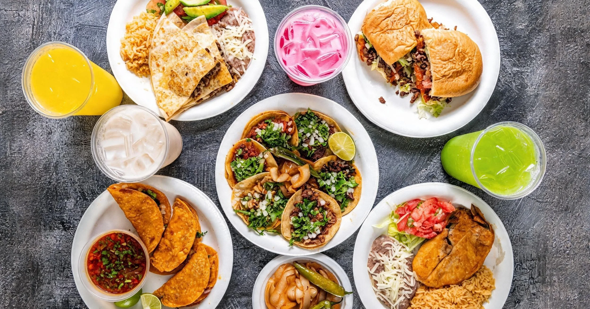Variety of Mexican dishes on plates and drinks on a dark surface, including tacos, burritos, fried fish, rice, and fruit-flavored drinks.