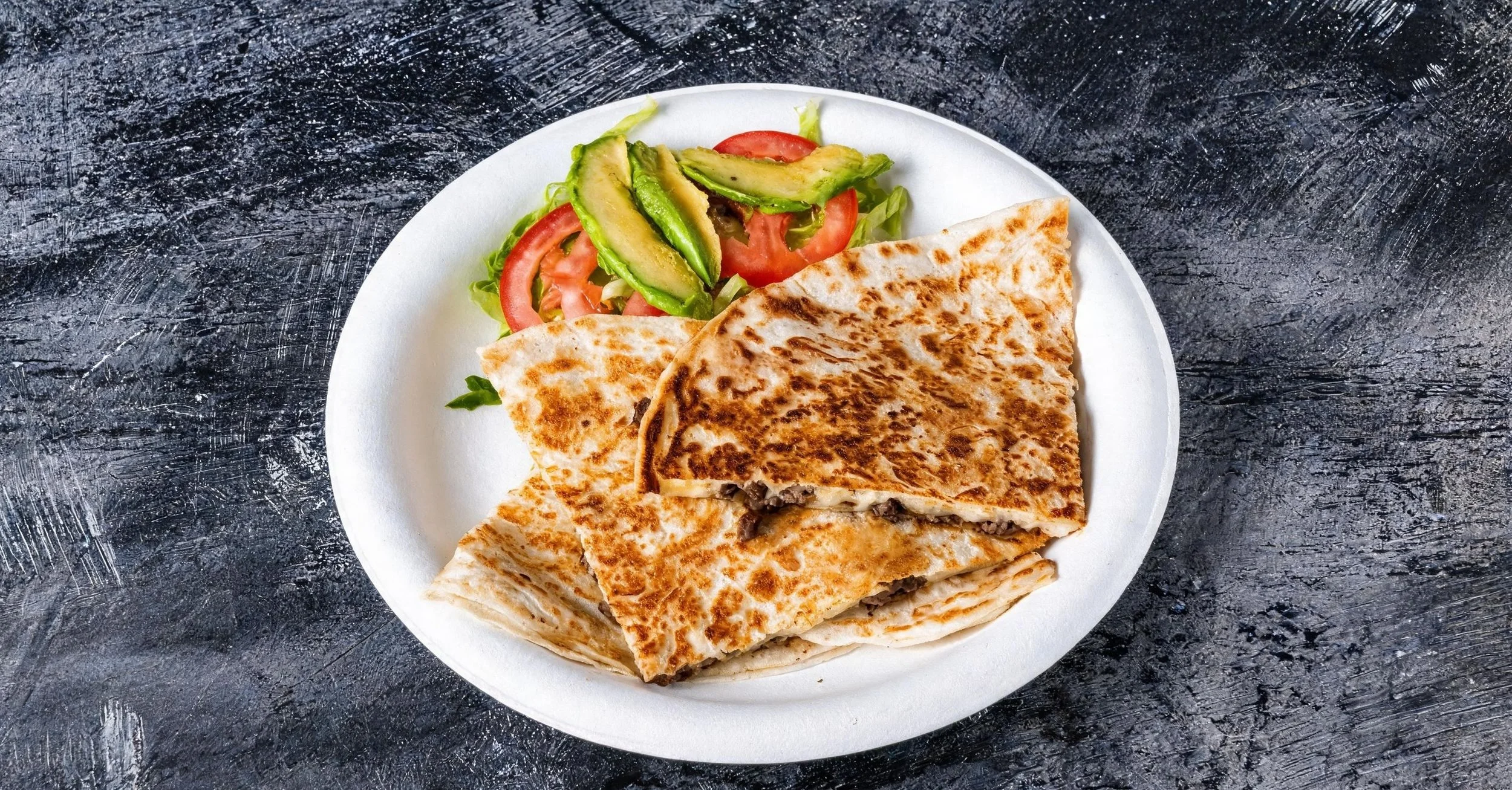 Plate with a quesadilla cut into quadrants and a side salad of sliced tomatoes, avocado, and lettuce, on a dark textured surface.