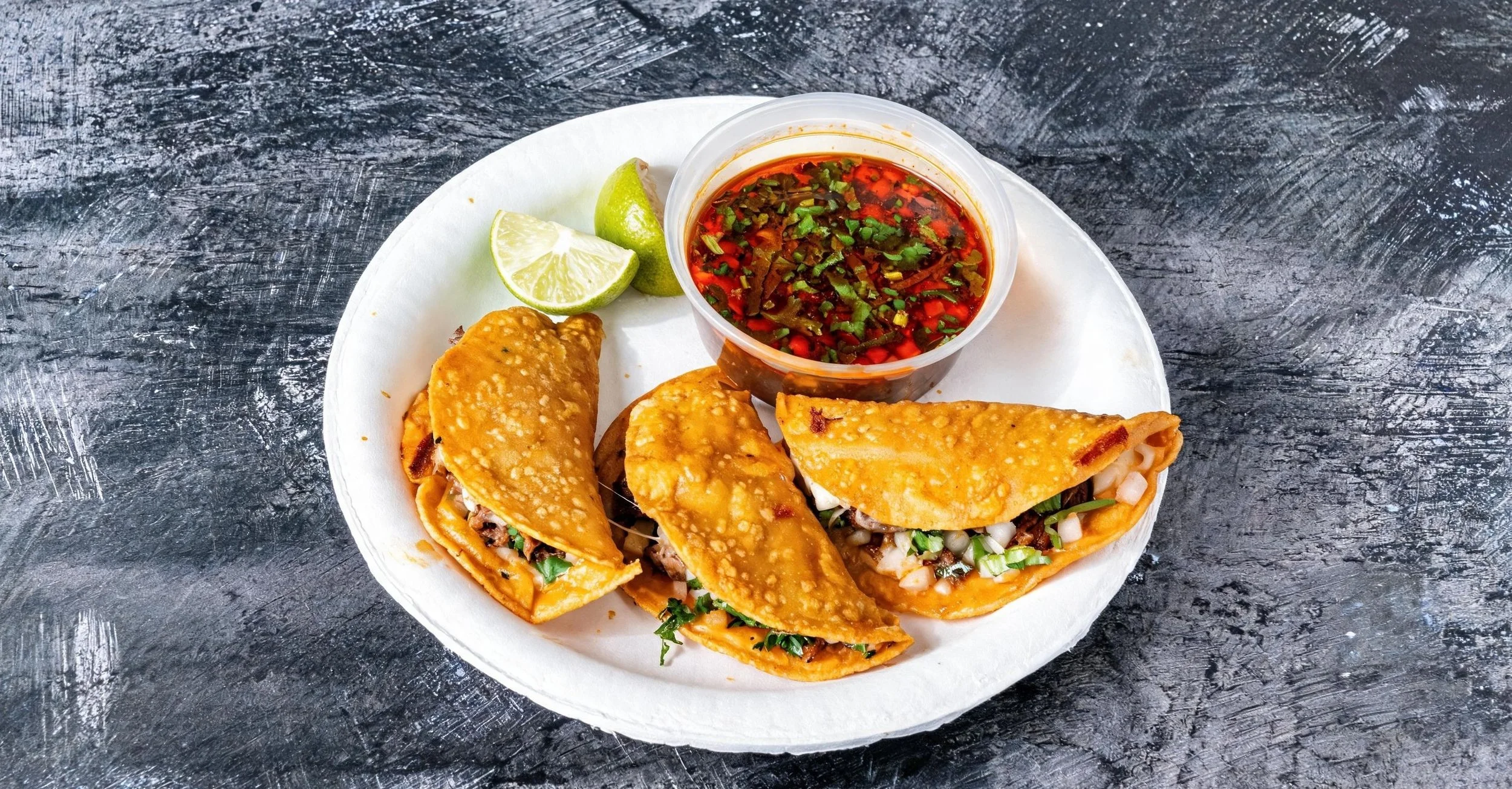 A white disposable plate holding three beef tacos with chopped onions and cilantro, accompanied by a small cup of red salsa and lime wedges, on a dark textured surface.