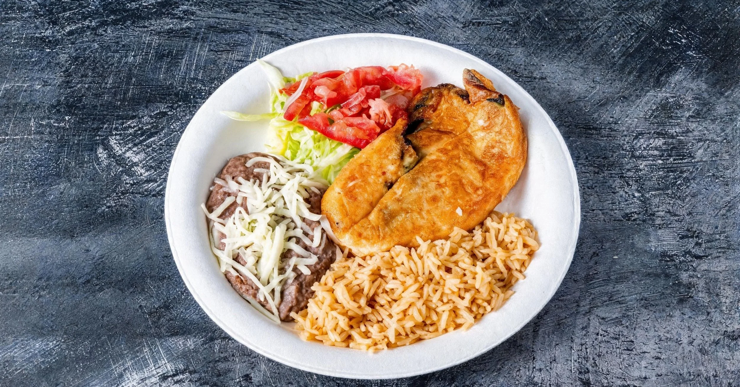 A white disposable plate with Mexican food including Mexican rice, refried beans topped with shredded cheese, a crispy quesadilla, lettuce and tomato salad, all on a dark textured surface.