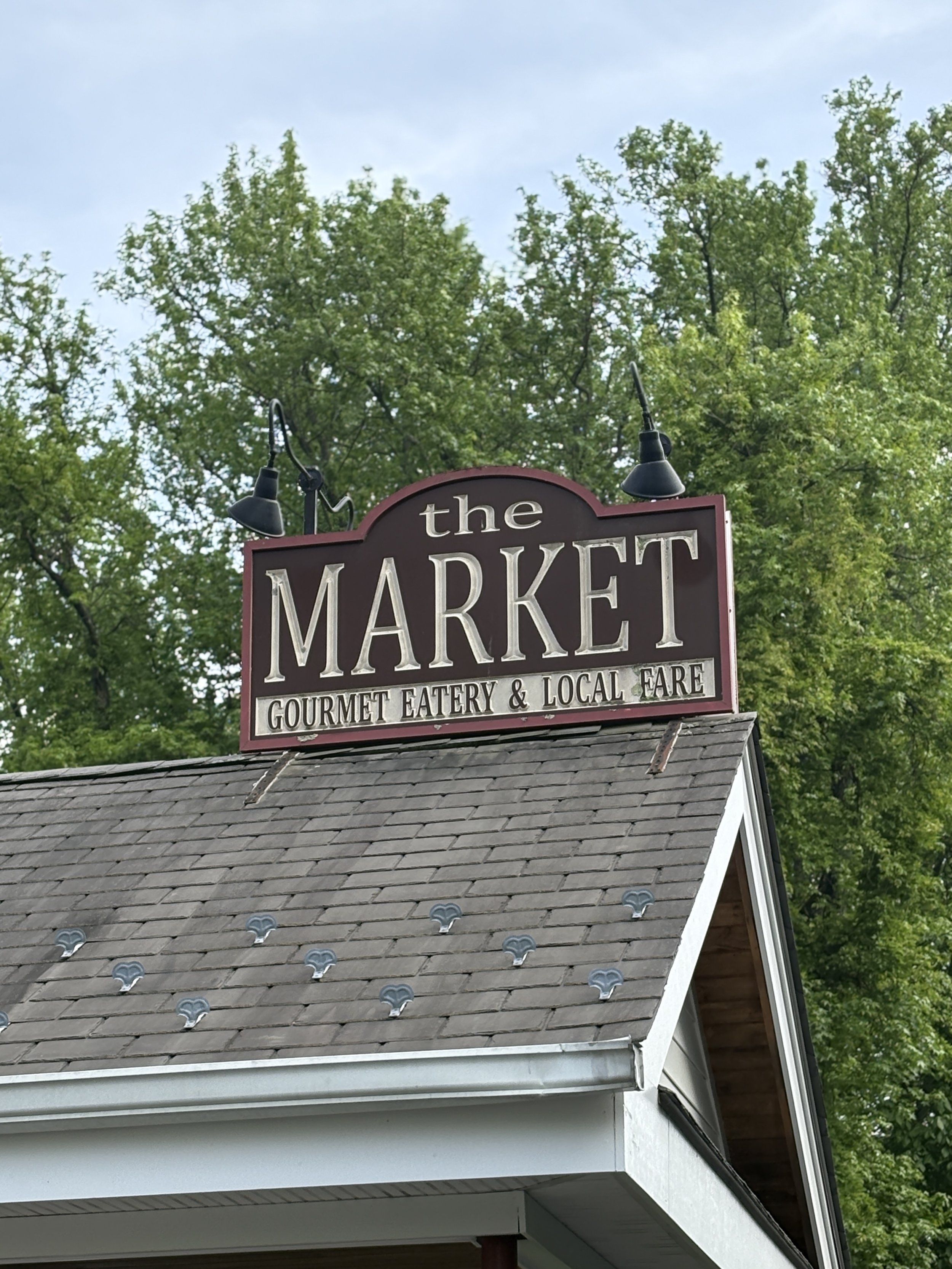 Sign reading 'the MARKET' with word 'GOURMET EATER & LOCAL FARE' below it, mounted above a building with gray shingle roof, black light fixtures above the sign, and green trees in the background.
