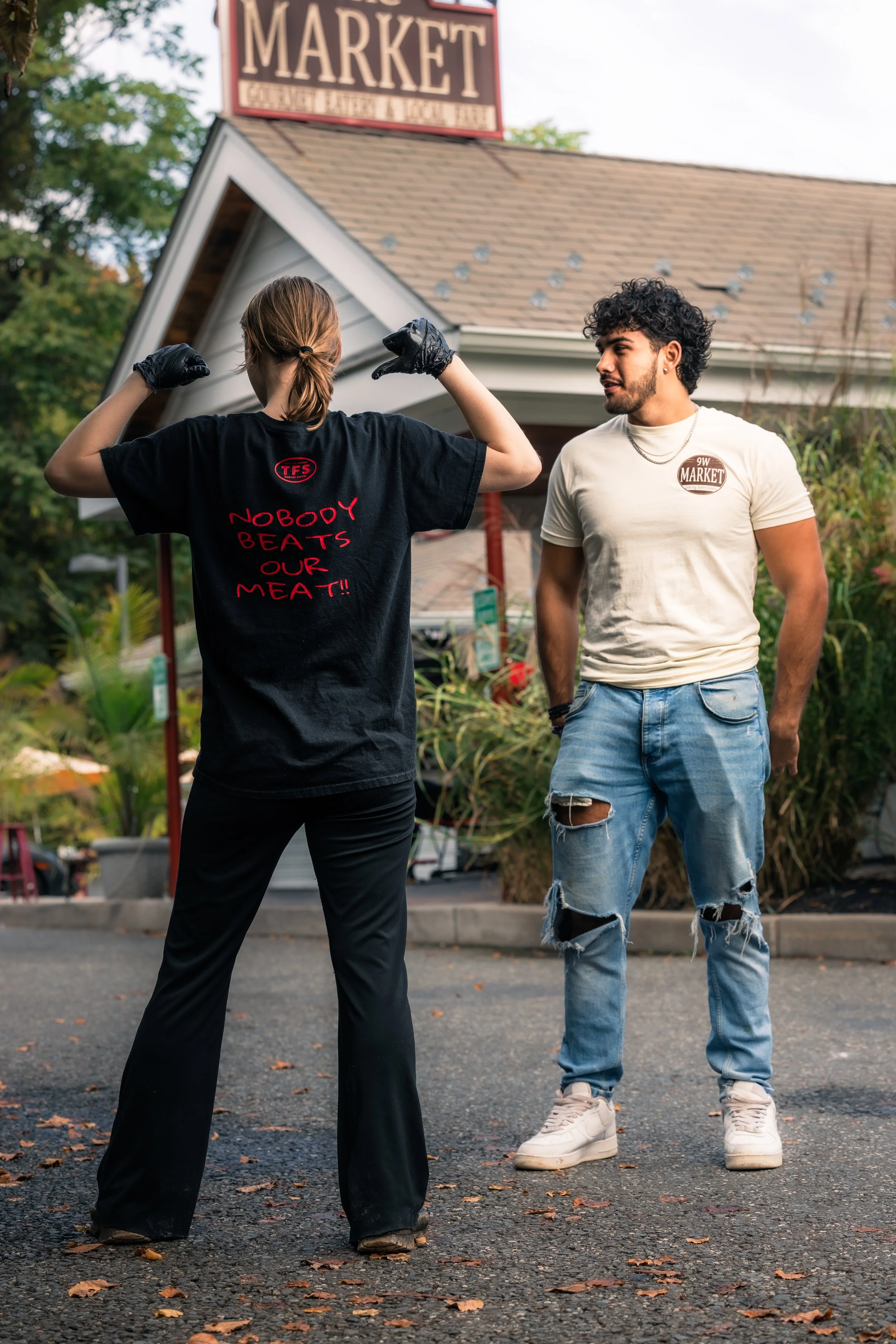 A woman flexing her arms displays a shirt that says 'Nobody Beats Our Meat!!' while a man stands nearby with his hands in his pockets outside a market store. The market sign is visible in the background.
