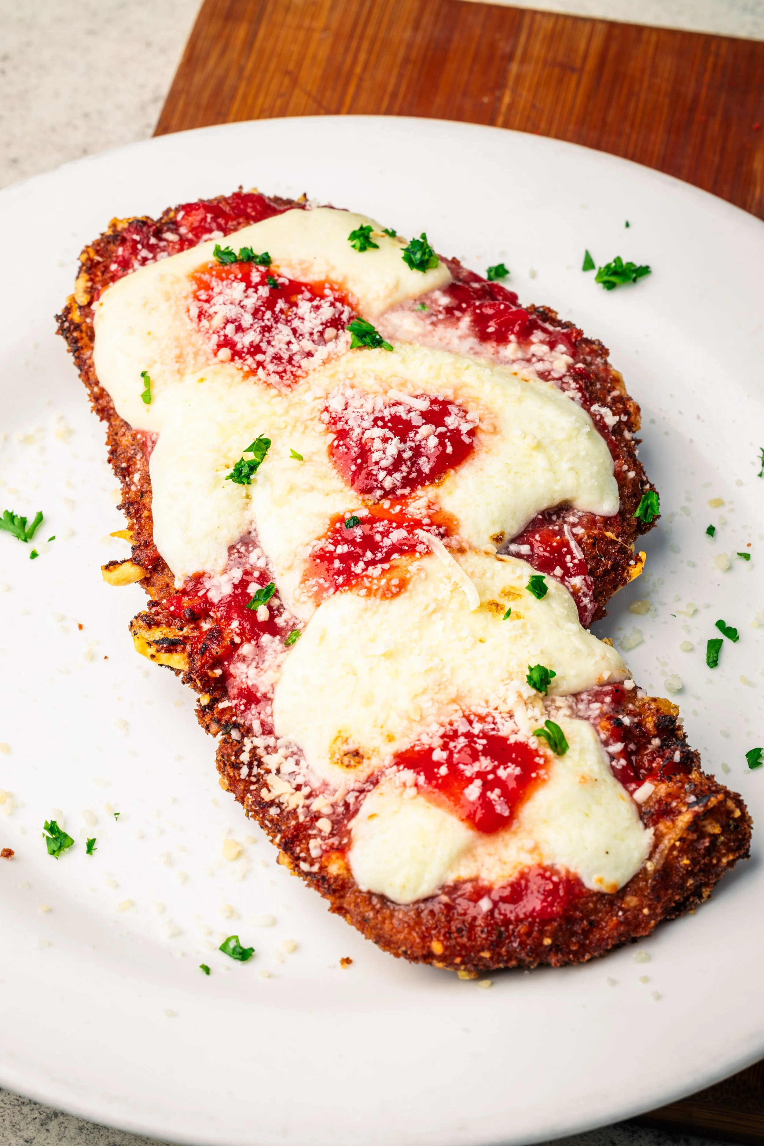 A breaded slice of eggplant topped with melted mozzarella cheese, marinara sauce, grated Parmesan cheese, and chopped fresh parsley on a white plate.
