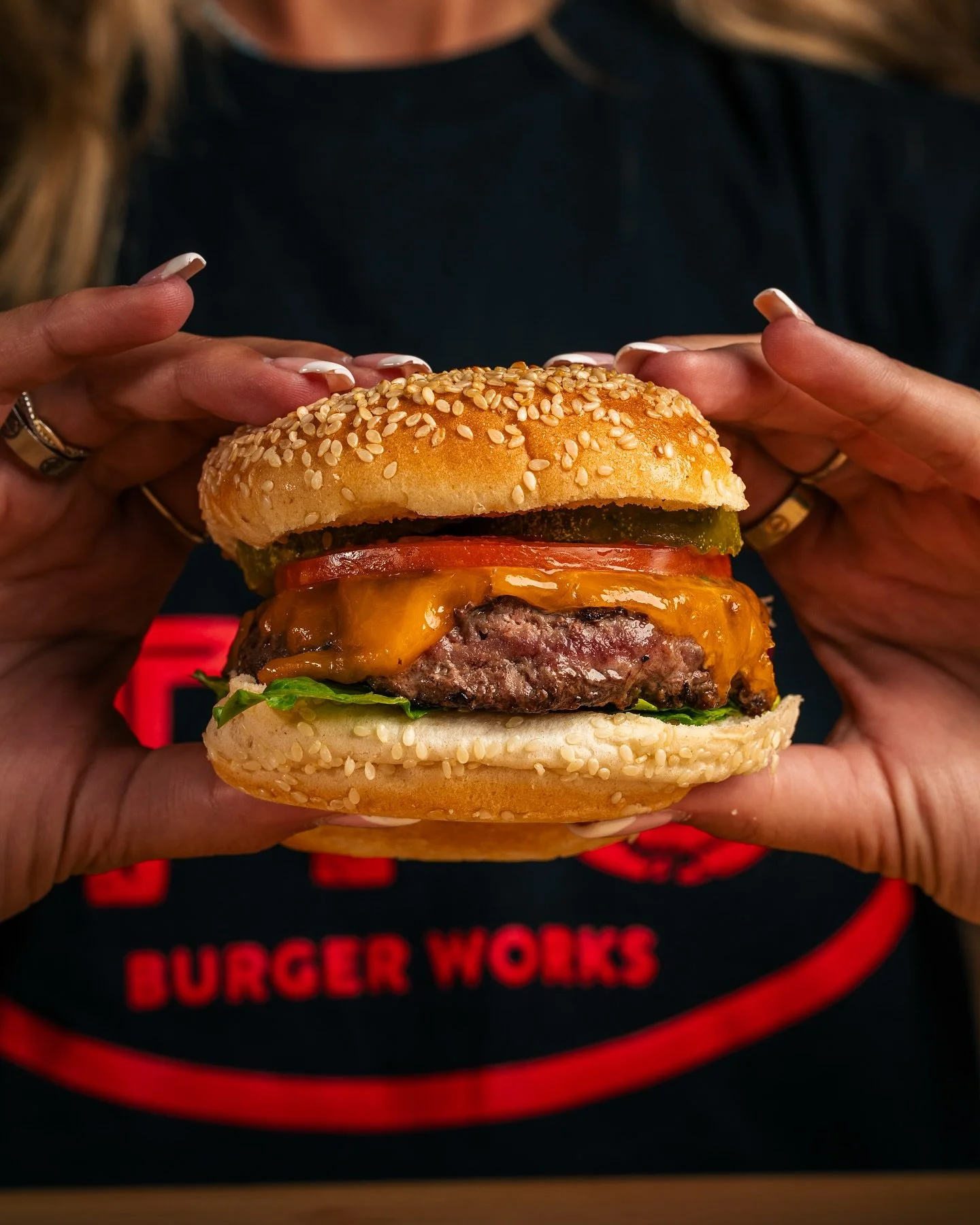 Person holding a cheeseburger with sesame seed bun, cheddar cheese, lettuce, tomato, pickles, and a beef patty, with a Burger Works t-shirt visible in the background.