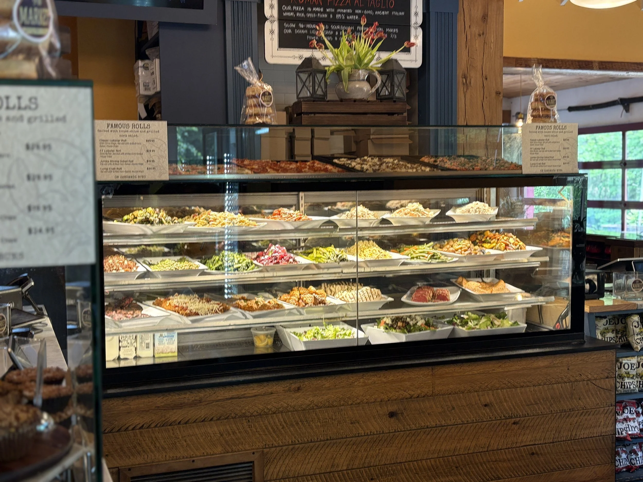 Display case with assorted salads and prepared foods inside a restaurant or deli, with a chalkboard menu and decorative flowers in the background.