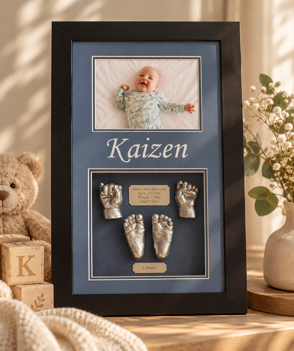 A framed baby keepsake with a smiling baby photo at the top, the name 'Kaizen' below, metallic baby hand and foot sculptures, a plaque with birth details, and a label indicating 6 weeks old.